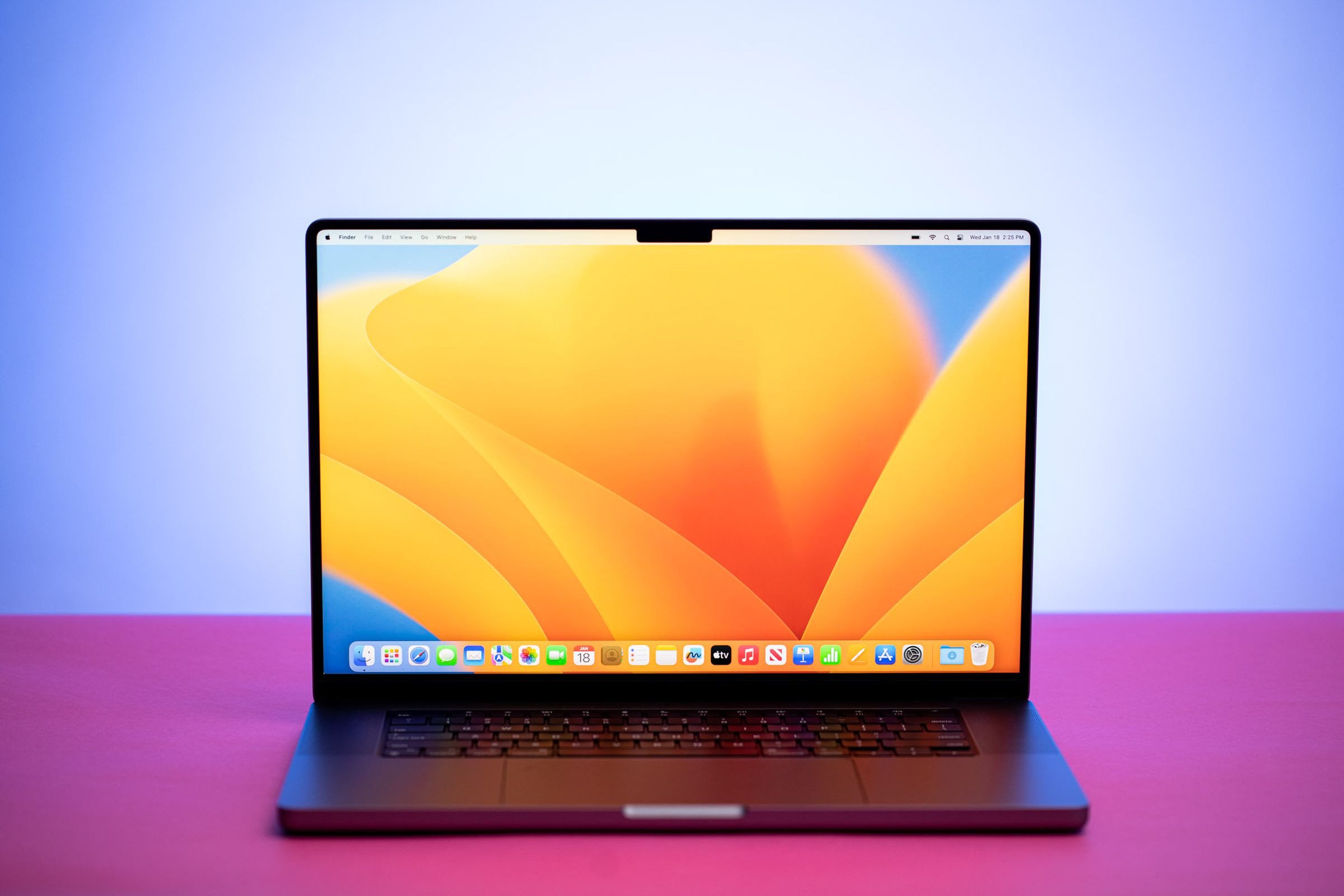 The MacBook Pro 16 (2023) on a pink table. The screen displays a blue and yellow desktop pattern.