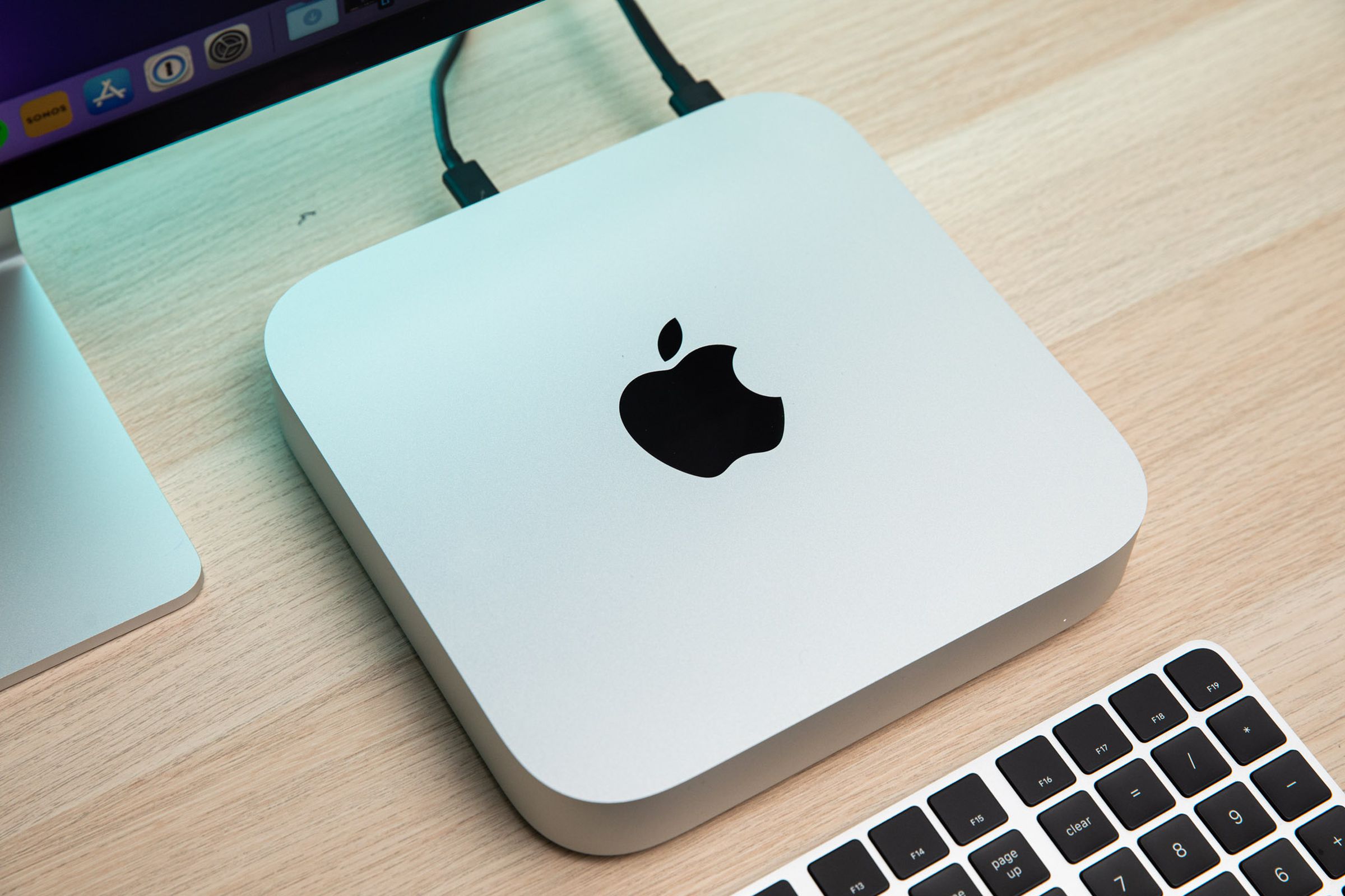 The Mac Mini sitting on a wood table underneath a monitor and beside a keyboard.