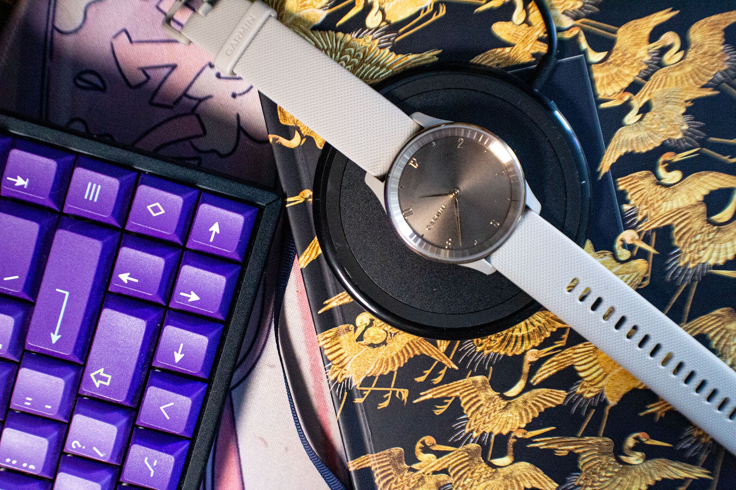 Garmin Vivomove Trend on a Qi charger, next to a purple keyboard and notebook with birds on it.