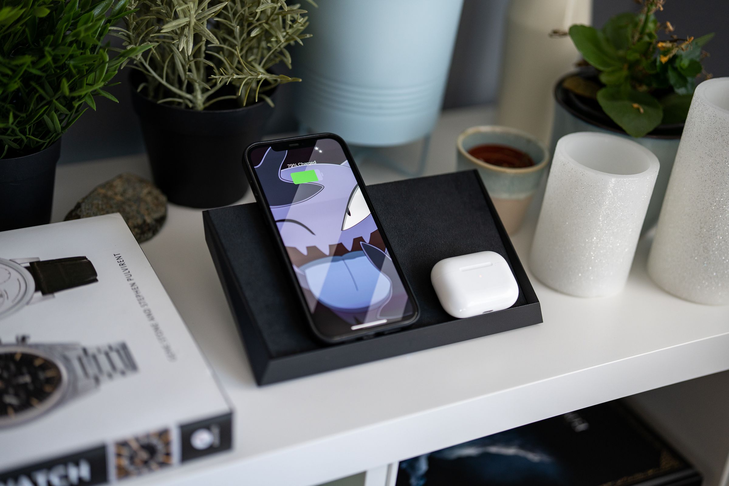 An iPhone and AirPods Pro case charging on Tesla’s wireless charging mat, sitting on a white end table.