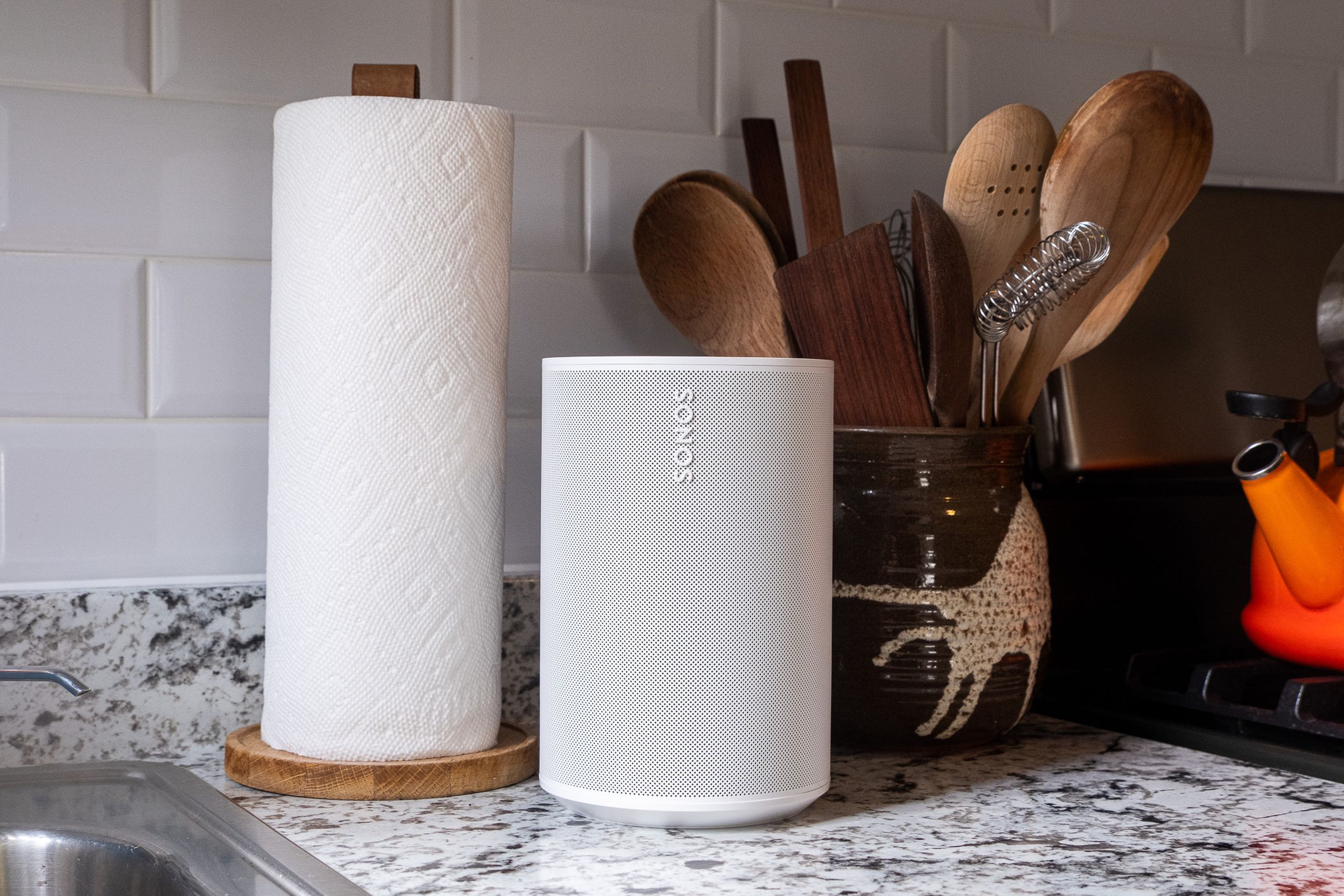 A white Sonos Era 100 speaker sitting on a marble kitchen countertop beside a paper towel roll and cooking utensils.