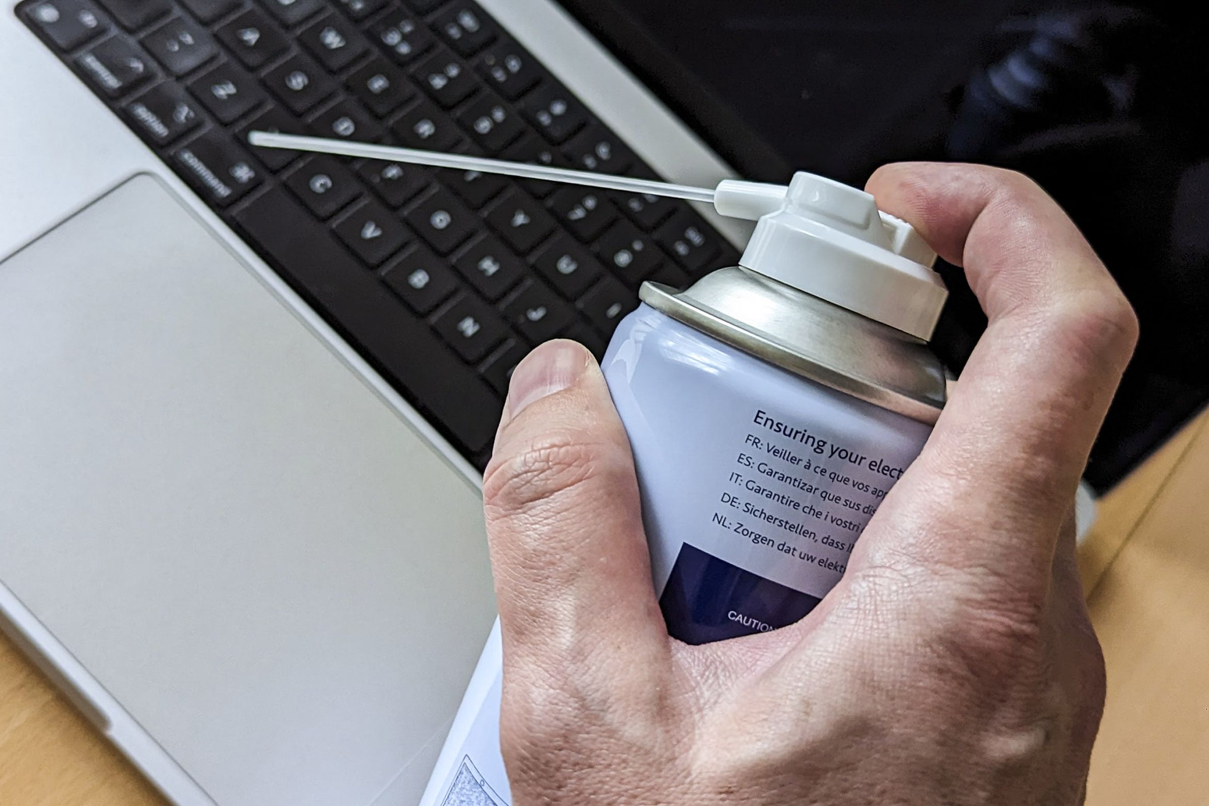 A hand holding a can of compressed air blowing into a laptop keyboard.