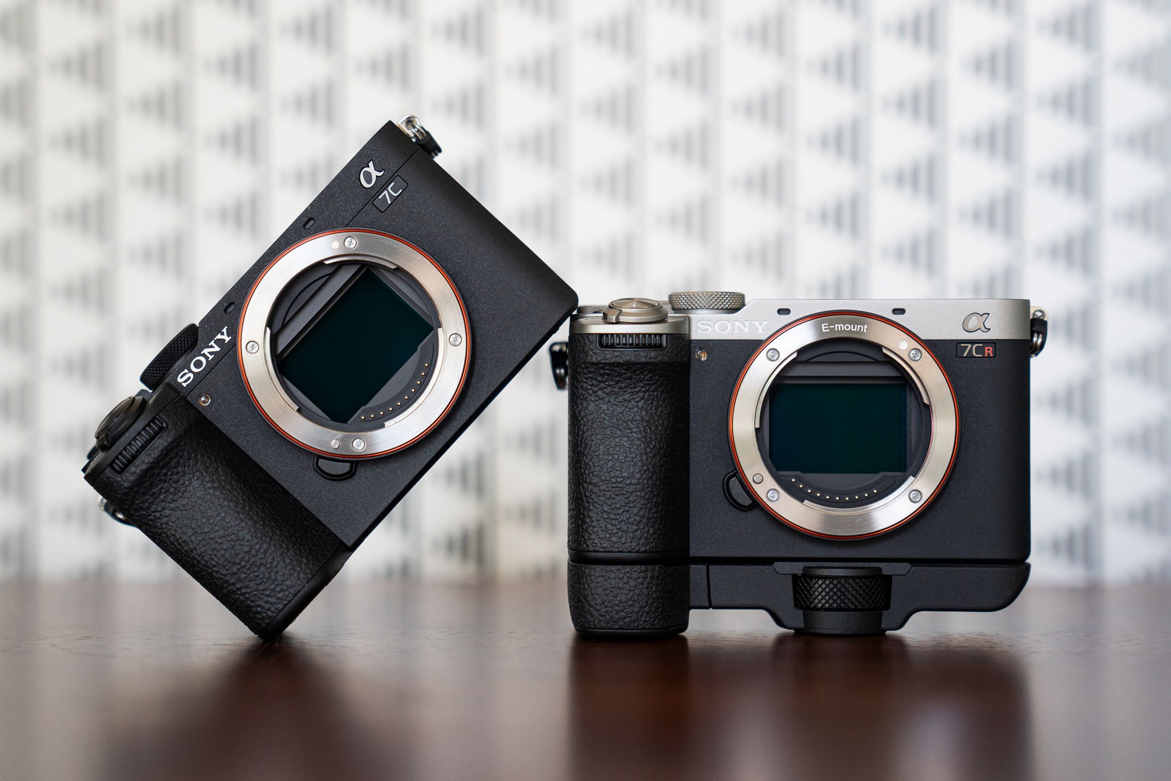 The Sony A7C II and A7C R cameras, sitting on a wood surface without lenses attached. The A7C II in black is angled and resting one side on the silver / black A7C R, which is mounted to its own handgrip.