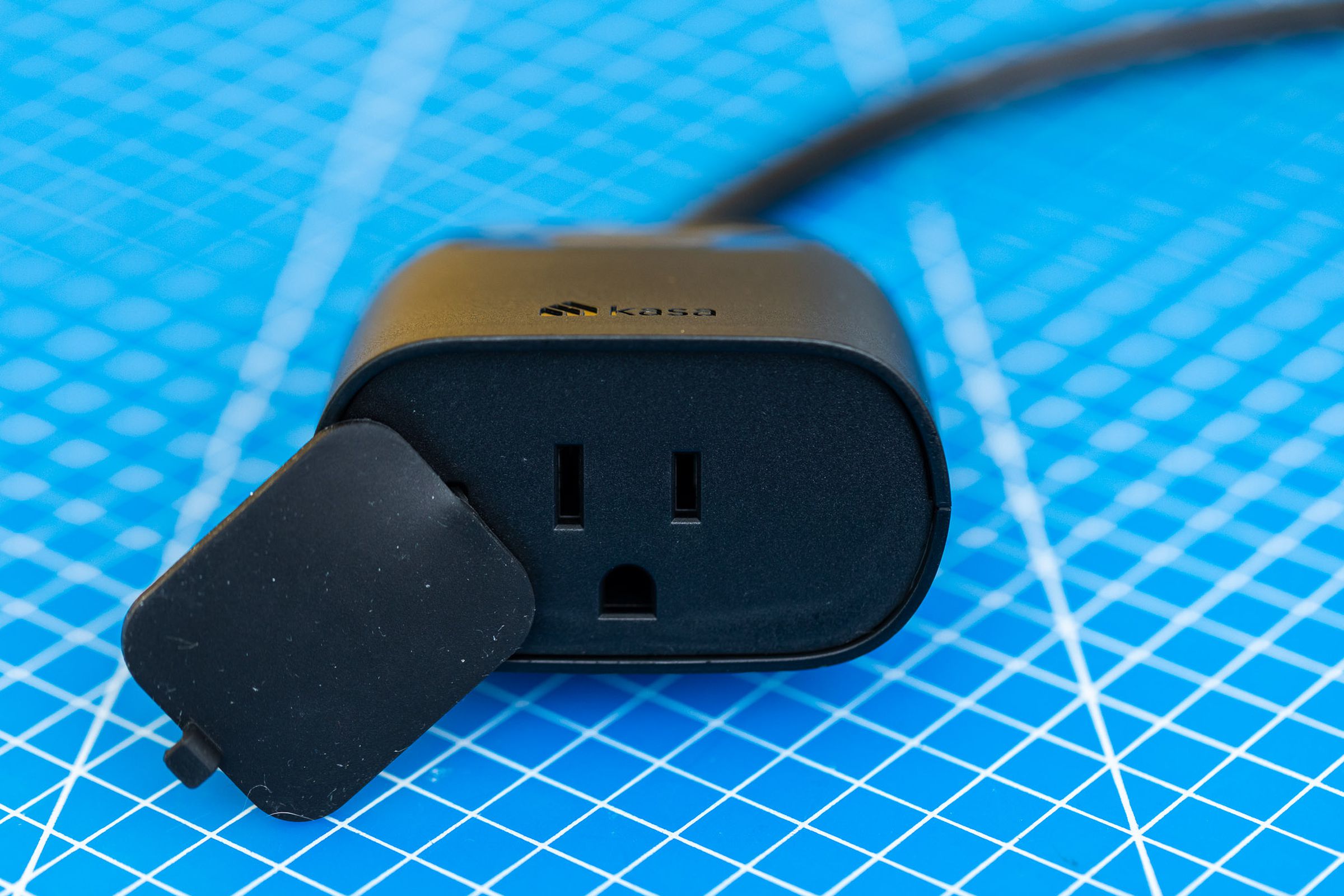 A black, two-outlet outdoor smart plug. It has a short cable leading to its own power plug, and a flap to protect the outlets from moisture. It is on a blue background with a grid of white lines.