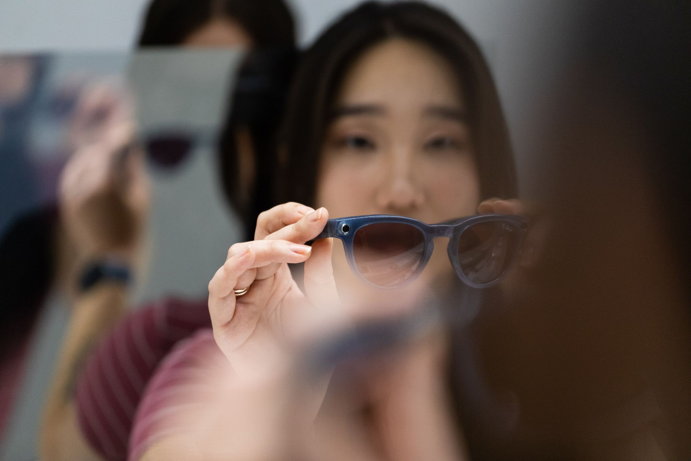 Person holding Ray-Ban Meta Smart Glasses in front of them while another person peers through a double-sided mirror in the background.