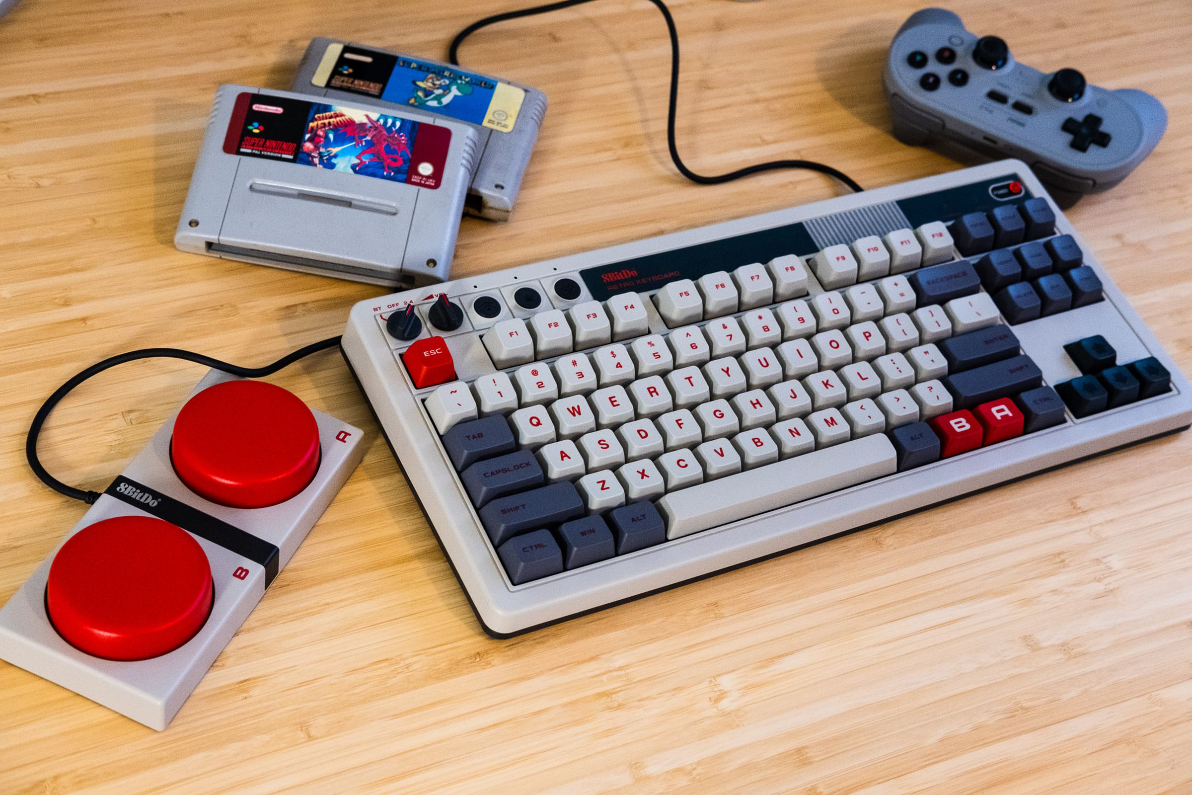 8BitDo Retro Keyboard on a desk next to NES-themed buttons, controller, and SNES cartridges.