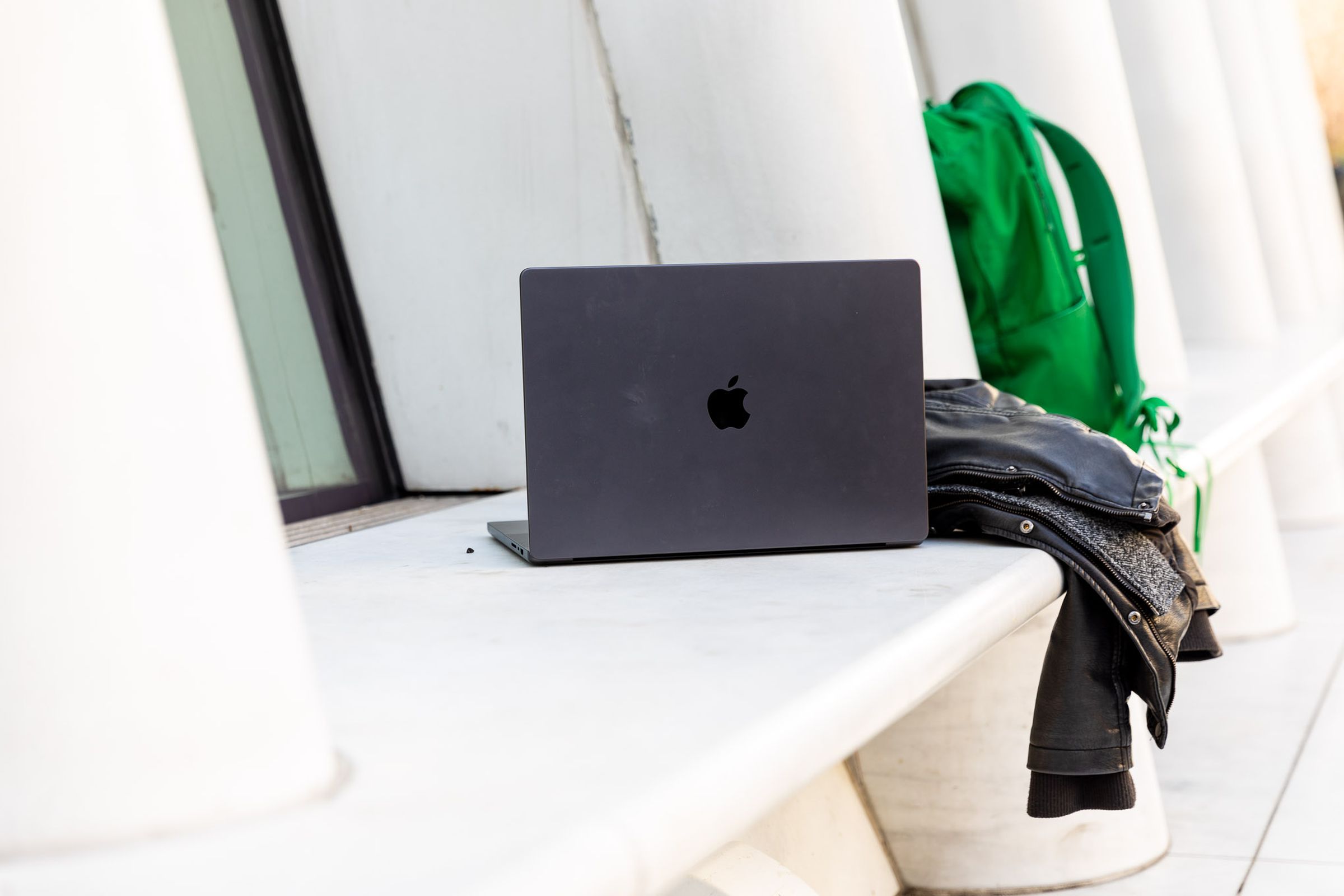 Wide view of the MacBook Pro 16 in space black’s cover, next to a leather jacket and green backpack with white columns in the background.