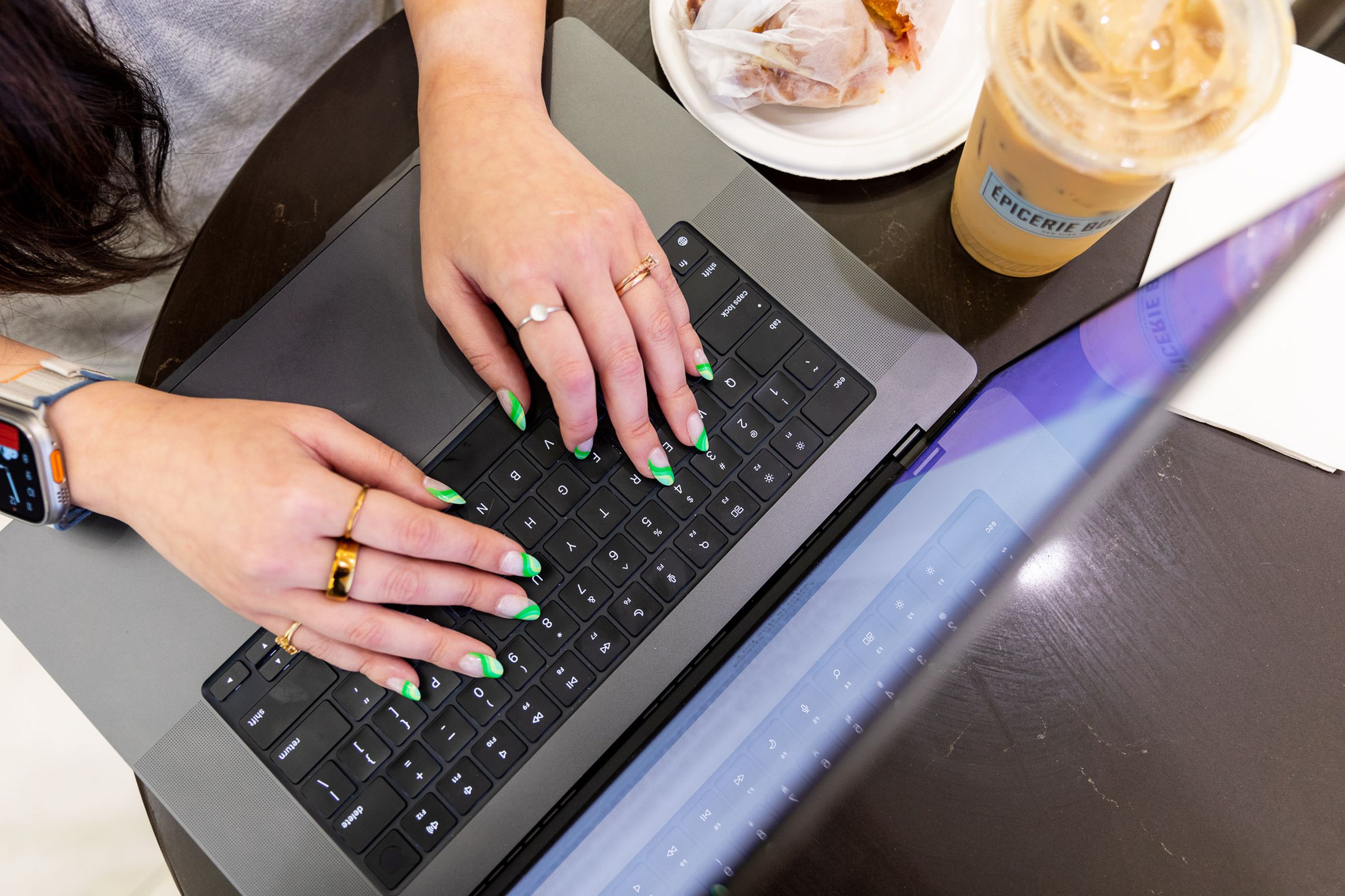 Top-down view of person with green nails typing on an M3 Max MacBook Pro 16, with a sandwich and coffee to the side. The corner of the laptop dangles off the edge.