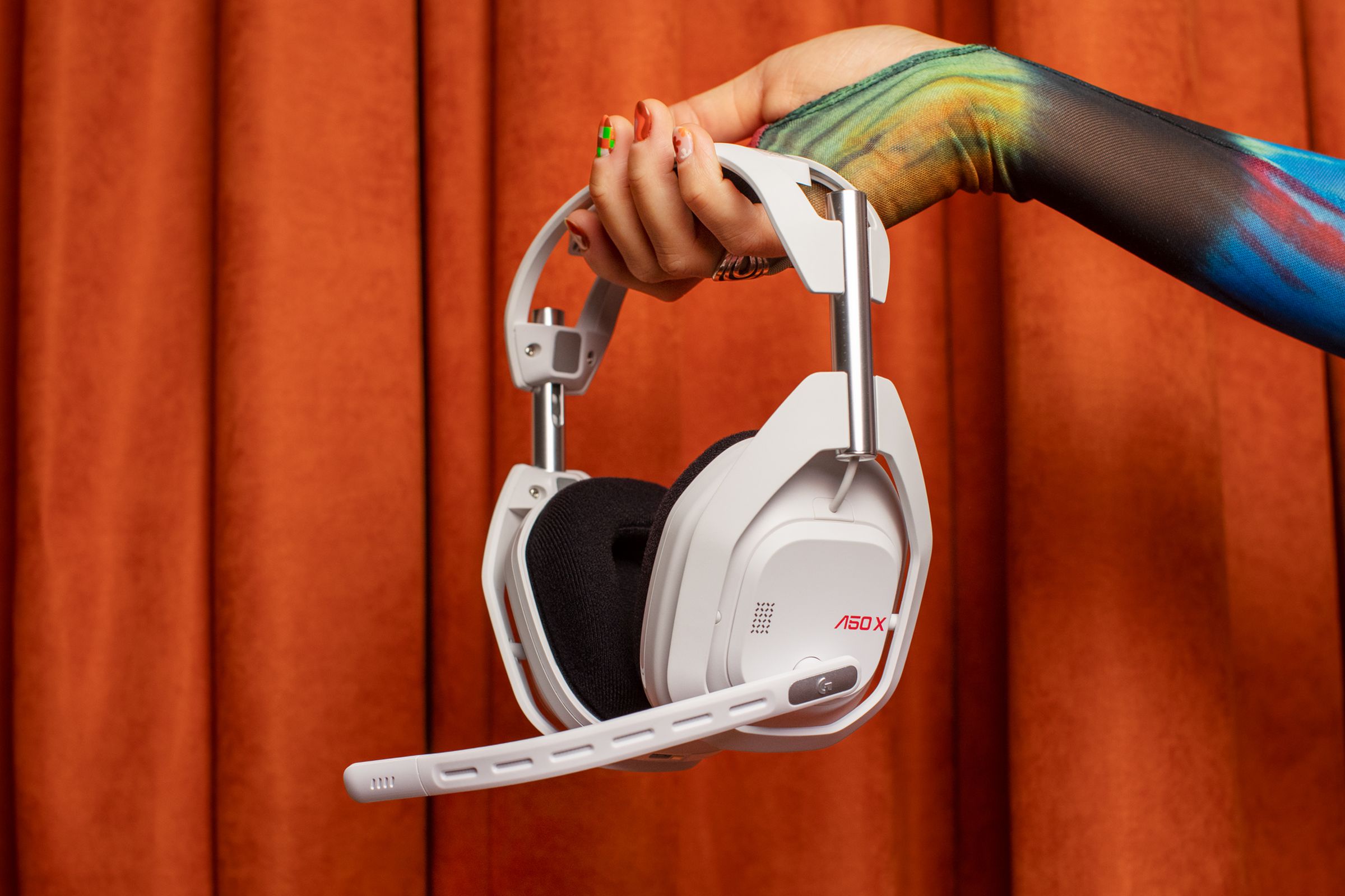 A person’s hand holding the white Astro A50 X gaming headset in front of a fabric backdrop.