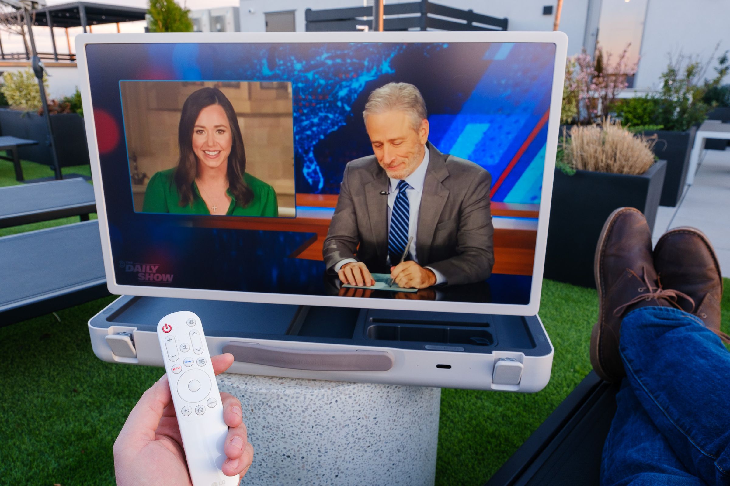 A photo of LG’s StanbyME Go briefcase TV being viewed on an apartment building rooftop with an episode of The Daily Show on the screen.