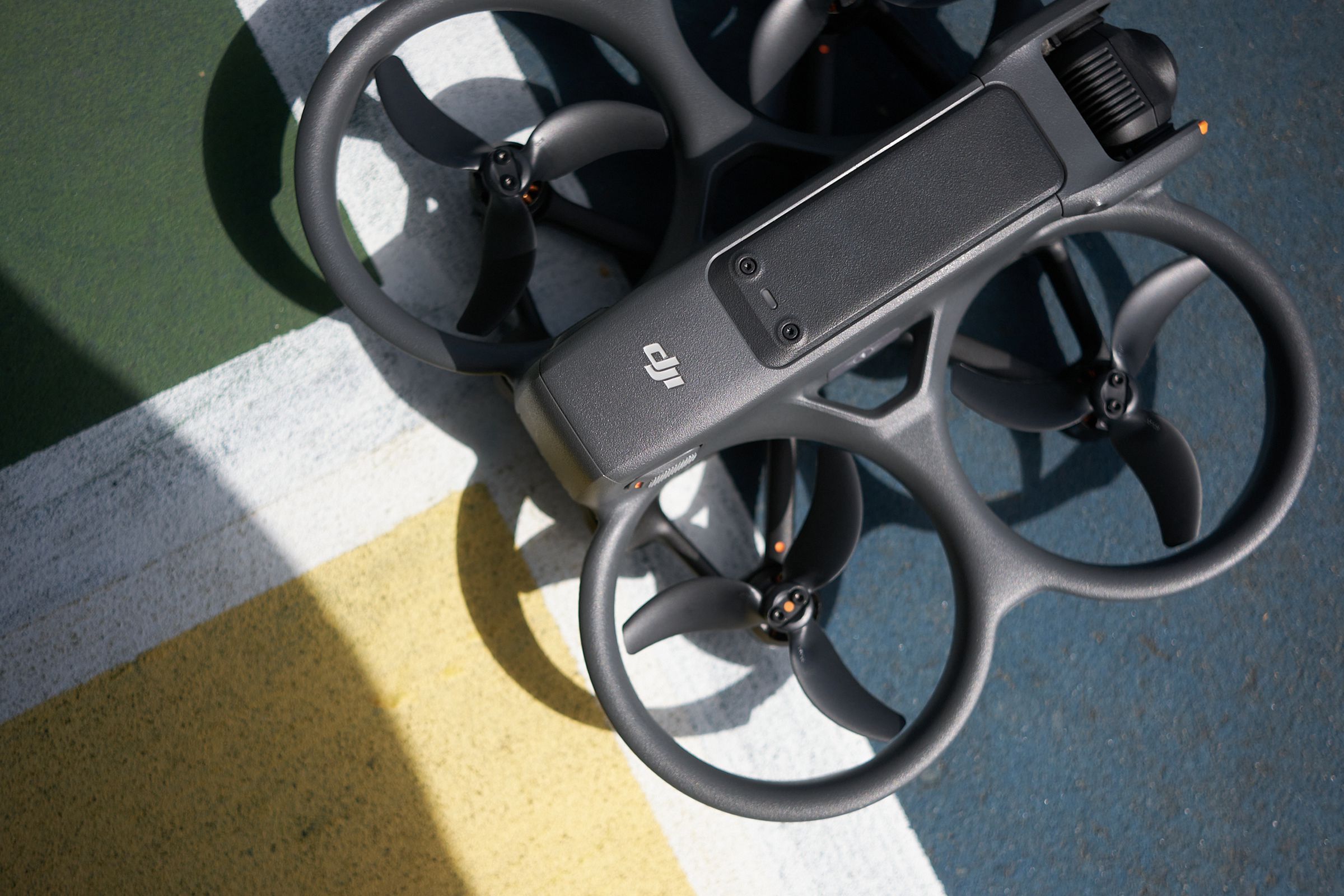 Three-quarters of the image are filled with the frame and three-bladed ducted propellors of a quadcopter drone with a slim gray body and a camera at one end.