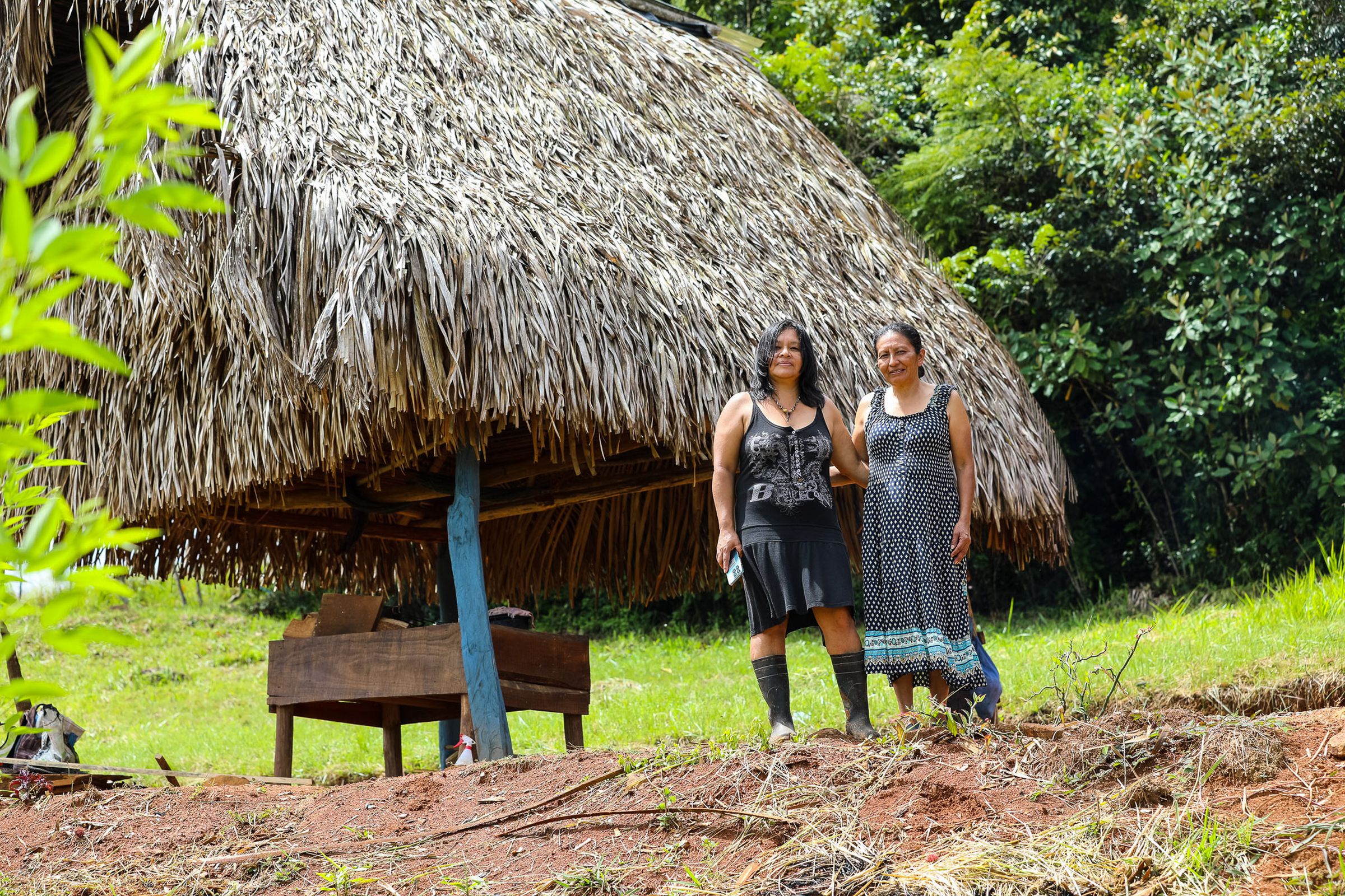 Two women stand and face the camera with their arms around each other. Behind them is an open shelter made of a thatched grass roof and wooden posts that still resemble tree trunks. 