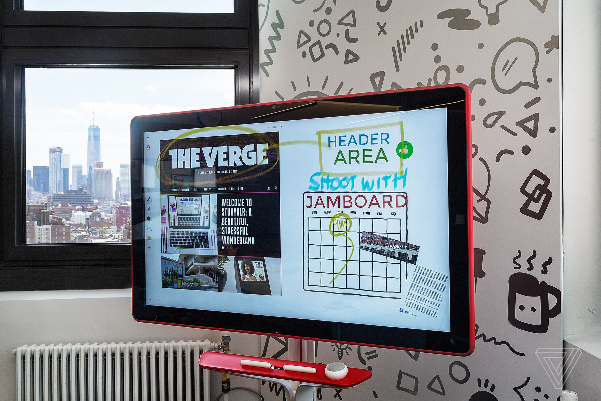 A Google Jamboard screen on a rod stand with styluses and the NYC skyline in the background outside the windows.