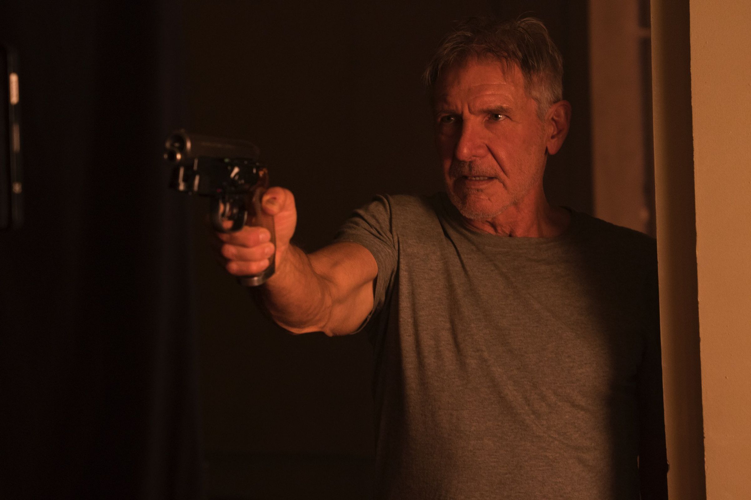Harrison Ford, as Deckard in Blade Runner 2049, stands in a doorway and points a gun.