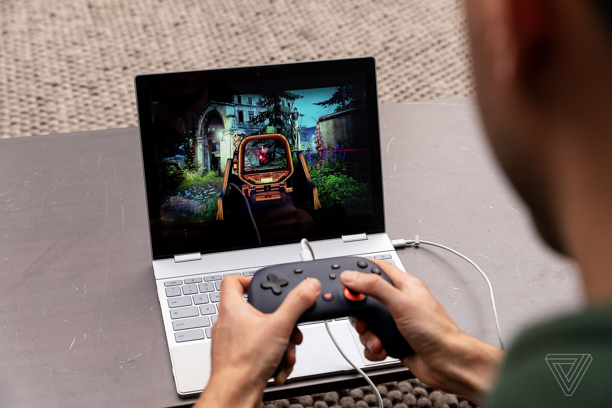 A person holds a Stadia controller in their hands. The controller is connected to a Chromebook, which is playing a game.