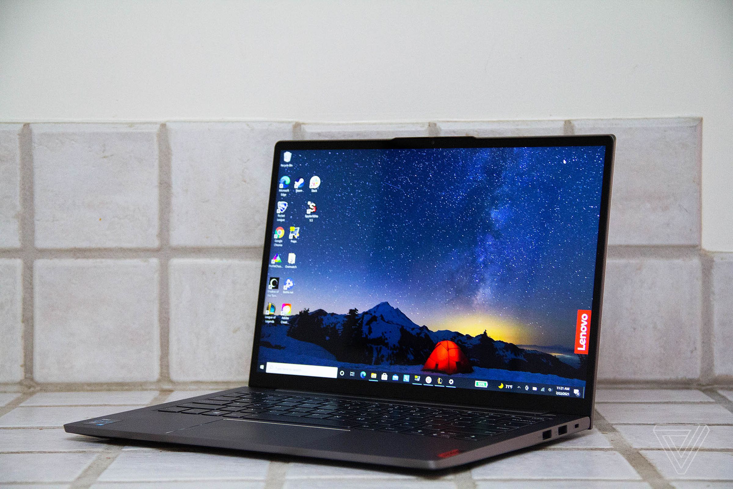 The Lenovo ThinkBook 13s on a white tiled counter, open, angled to the left. The screen displays a night mountain scene with a red tent in the center and the red Lenovo banner on the right side.