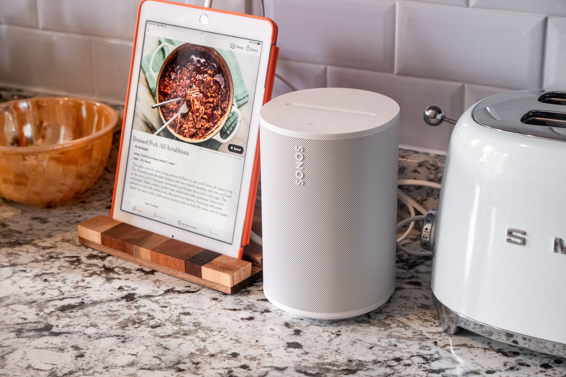 A photo of the Sonos Era 100 on a kitchen counter beside a toaster and iPad.