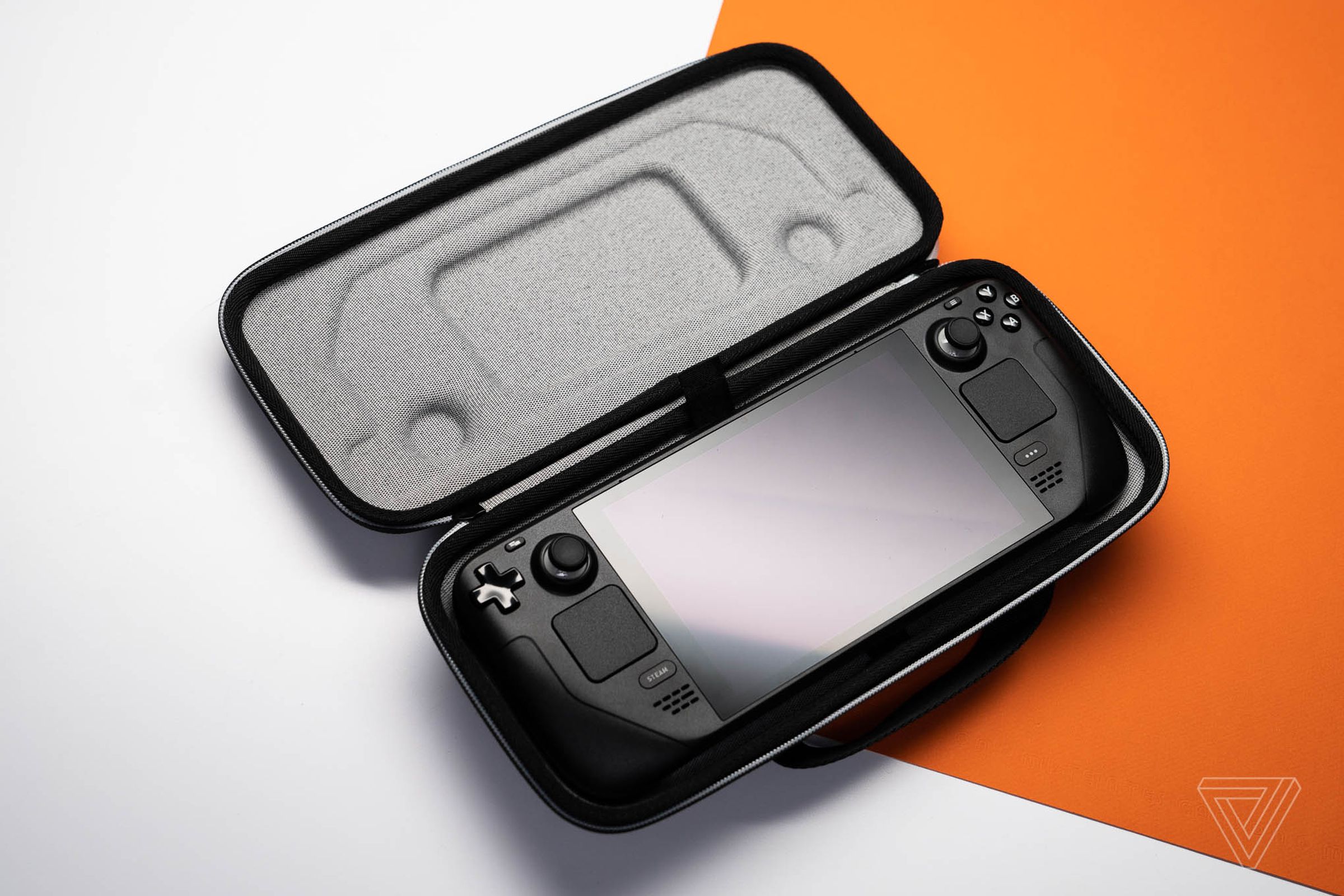A black gaming handheld with twin sticks and touchpads sits in an open grey zippered fabric case atop an orange and white background