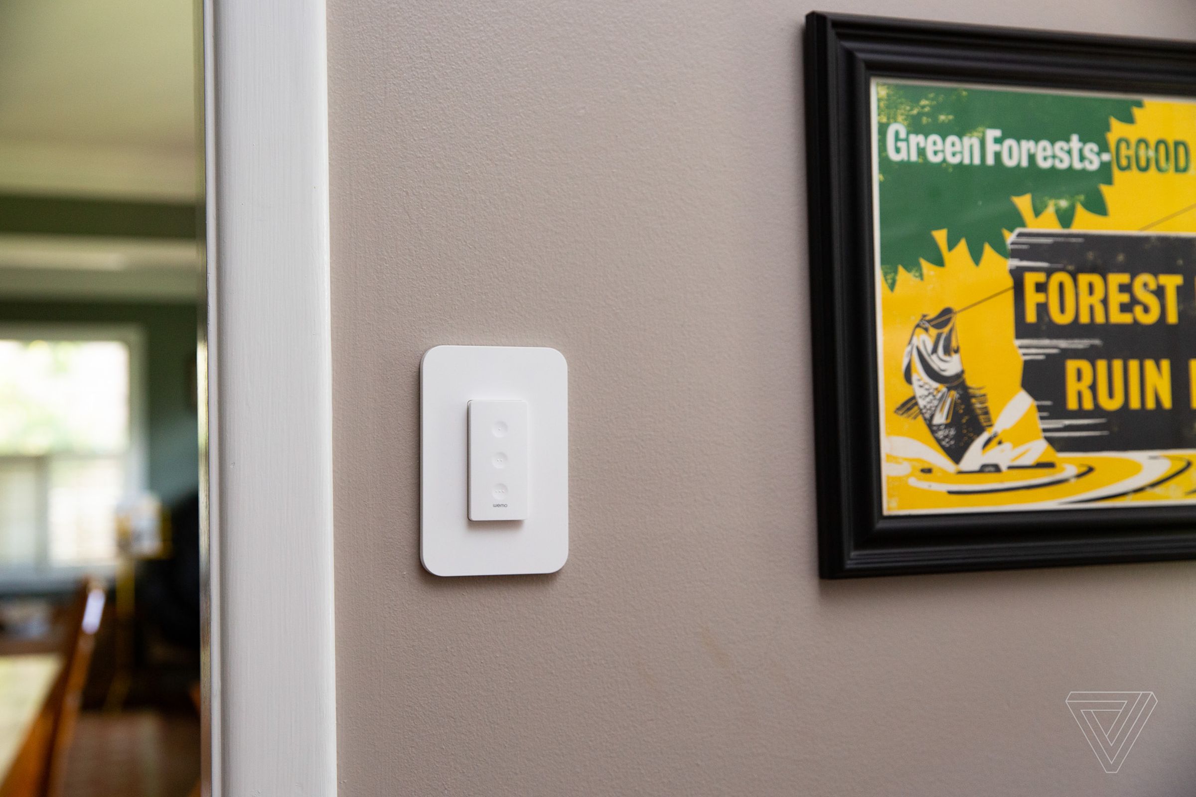 An image of a white light switch with three buttons. It is mounted on a tan wall. Green and yellow artwork in a black frame is also visible.