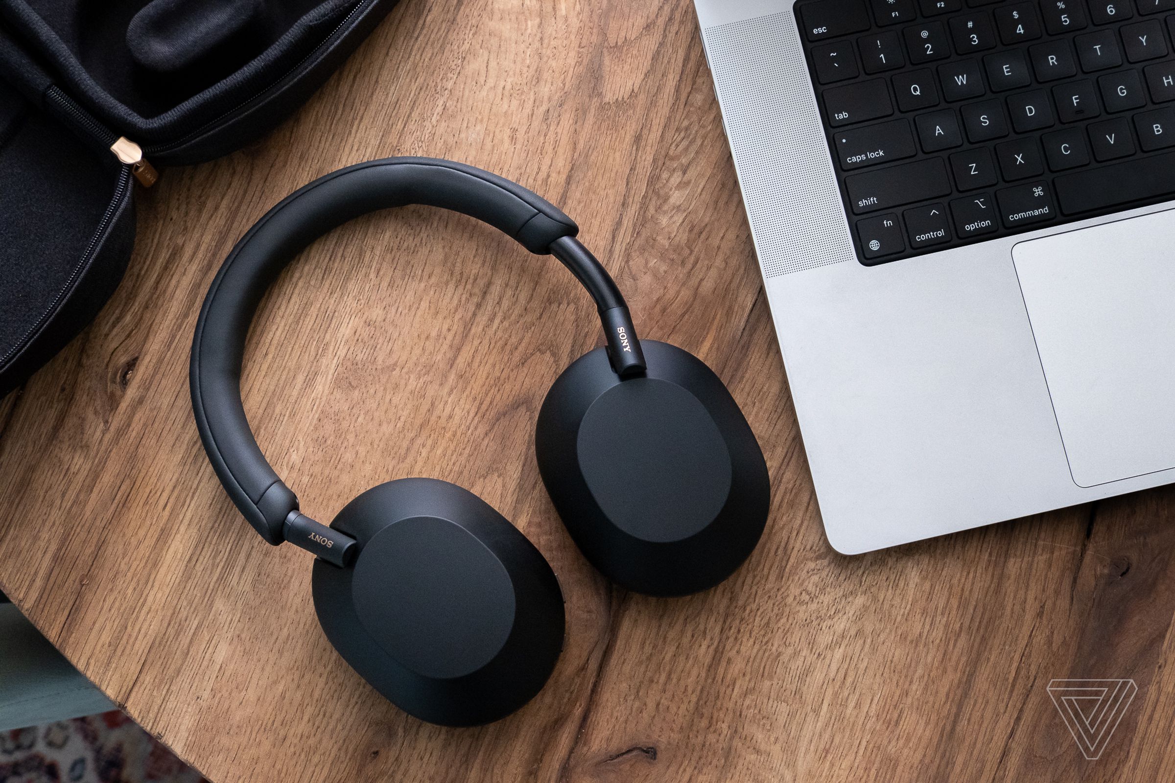 A pair of Sony WH-1000XM5 headphones, in black, resting flat on a wood table beside a laptop.