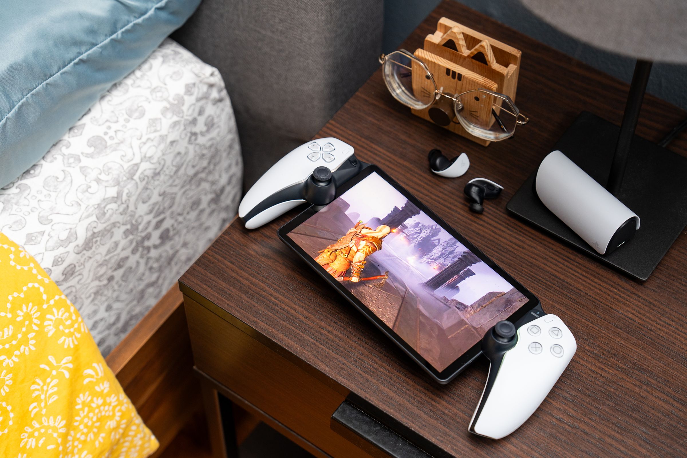 The PlayStation Portal sitting on a bedside table with a pair of earbuds. The handheld gaming device is streaming God of War: Ragnar?k off a PlayStation 5.