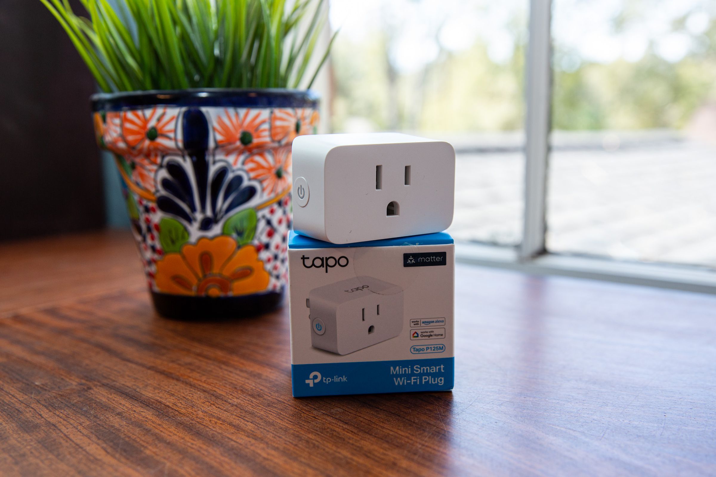 A smart plug on a box on a table by a window with a flower pot behind it.