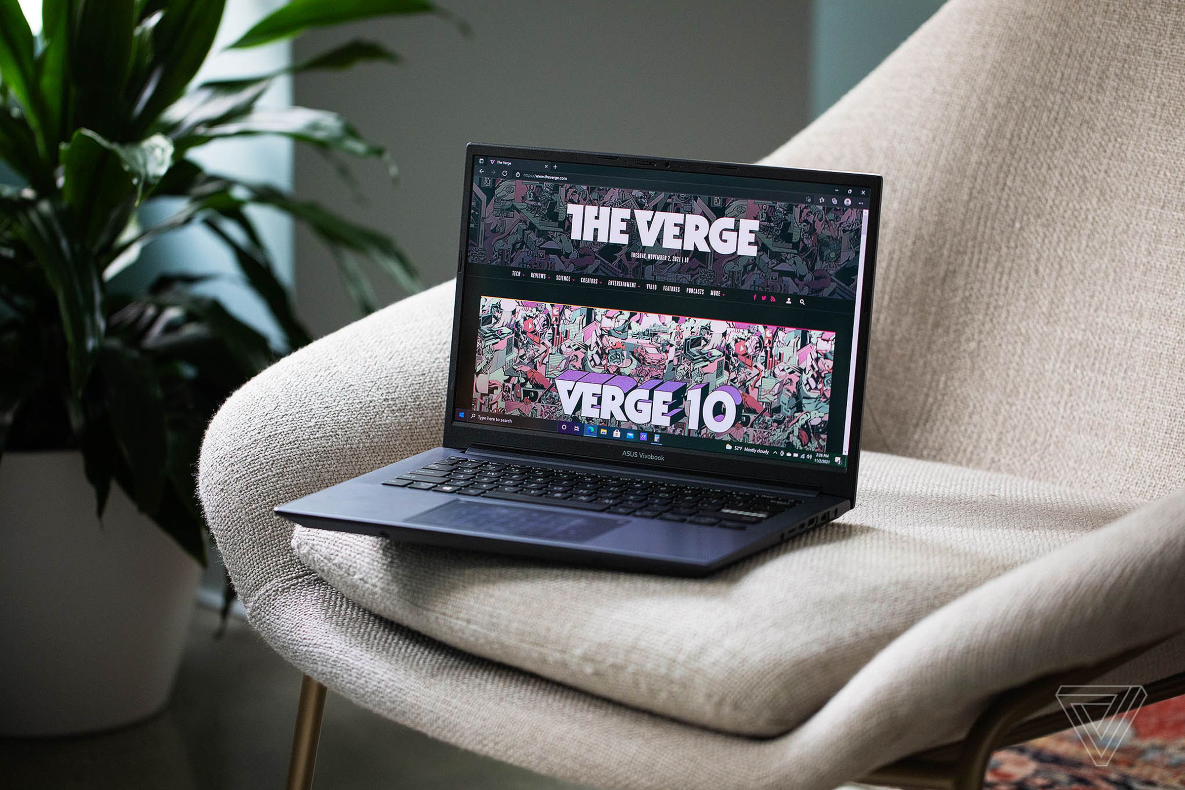 The Asus Vivobook Pro 14 open and angled to the left on a white plush chair with a houseplant to the left. The screen displays The Verge homepage.