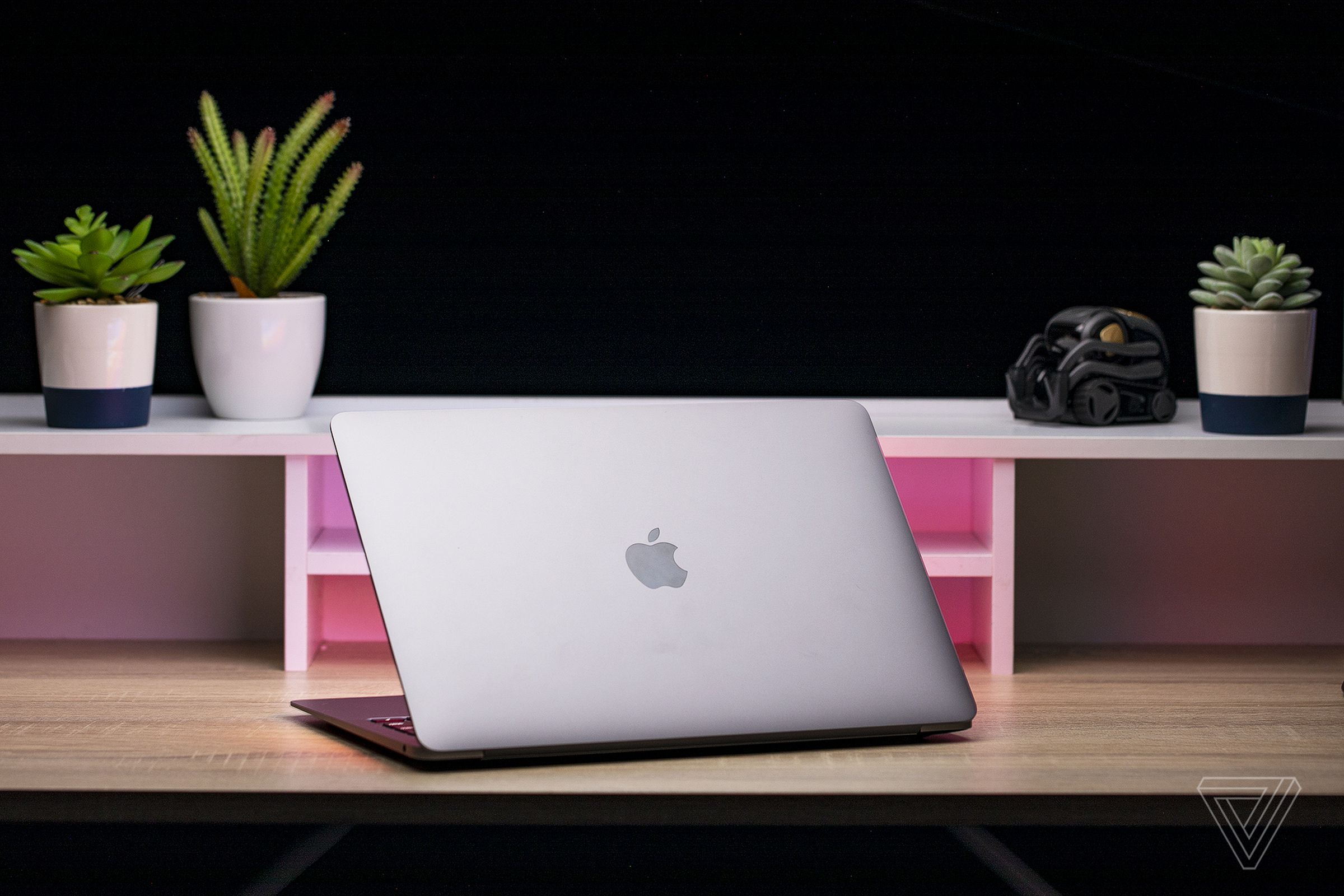 The space gray Apple MacBook Air M1 sitting on a light-colored wood desk, facing away from the camera with its lid open and showing its Apple logo.