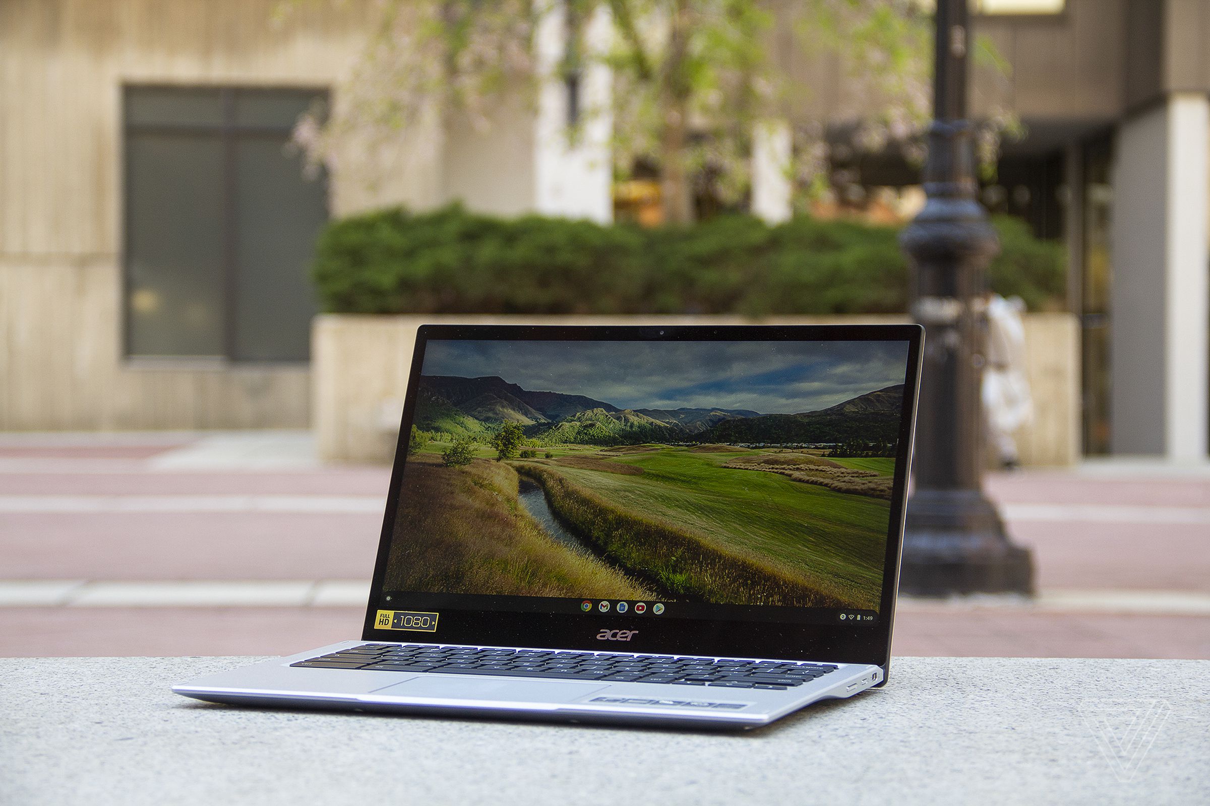 The Acer Chromebook Spin 513 sits on a stone bench, open, angled to the left. The screen displays a pastoral scene.