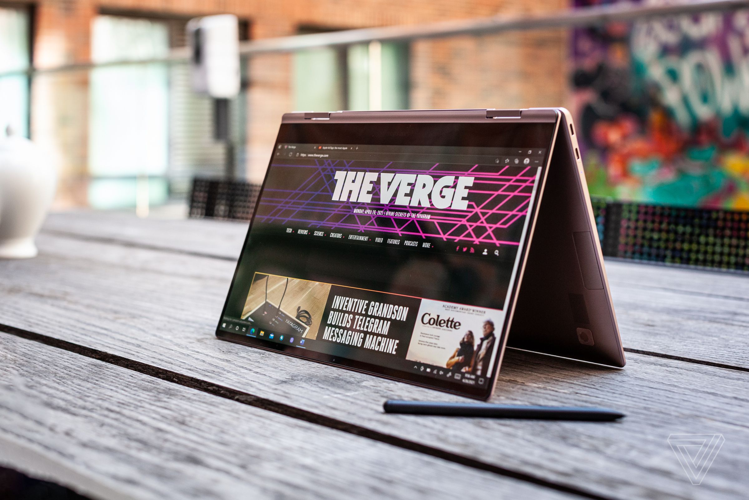 The Samsung Galaxy Book Pro 360 in tent mode, angled to the left, on an outdoor picnic table. The screen displays The Verge homepage. The S-Pen sits by its bottom right corner.