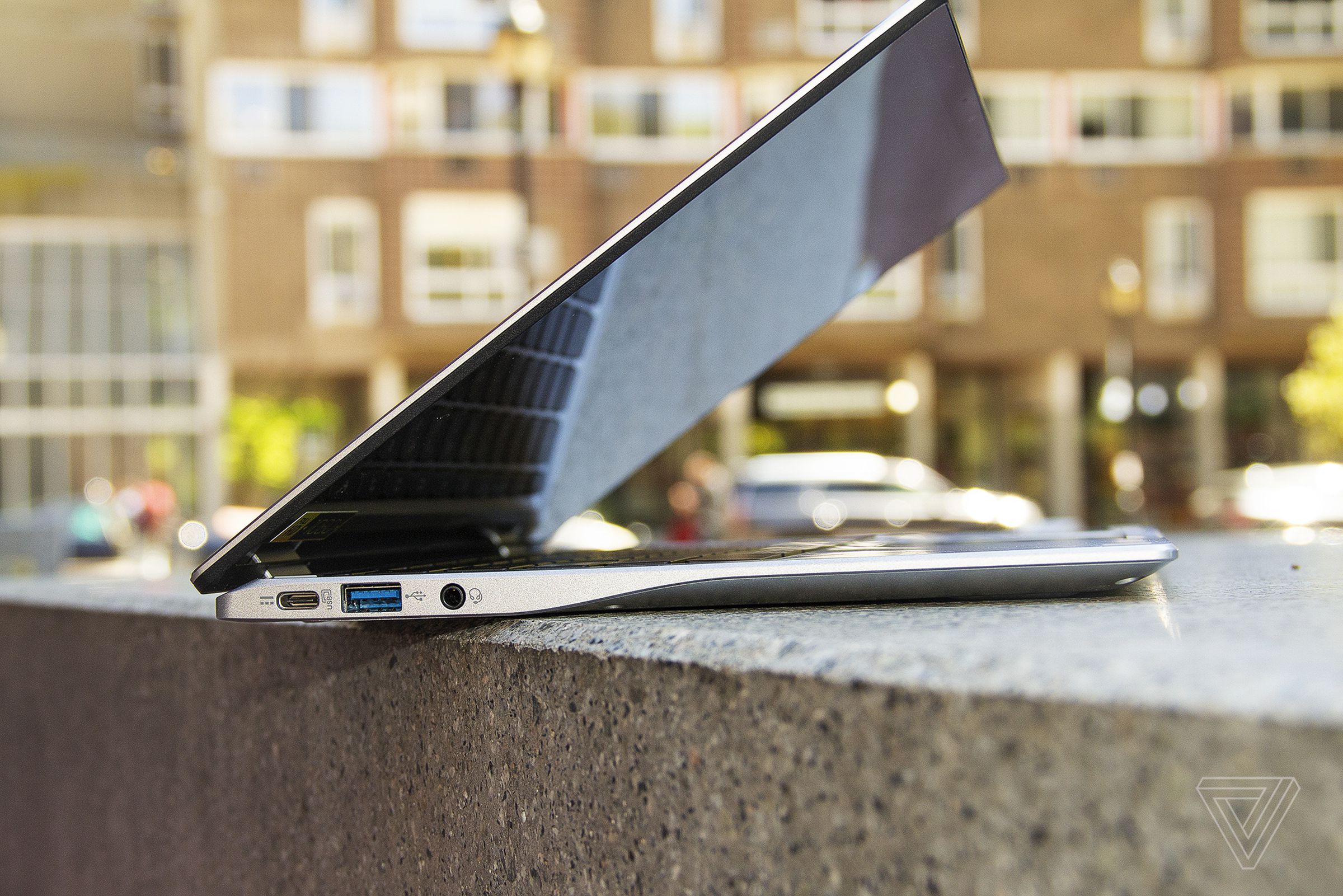 The Acer Chromebook Spin 513 seen from the left, half open, on a stone bench. An apartment building is in the background.