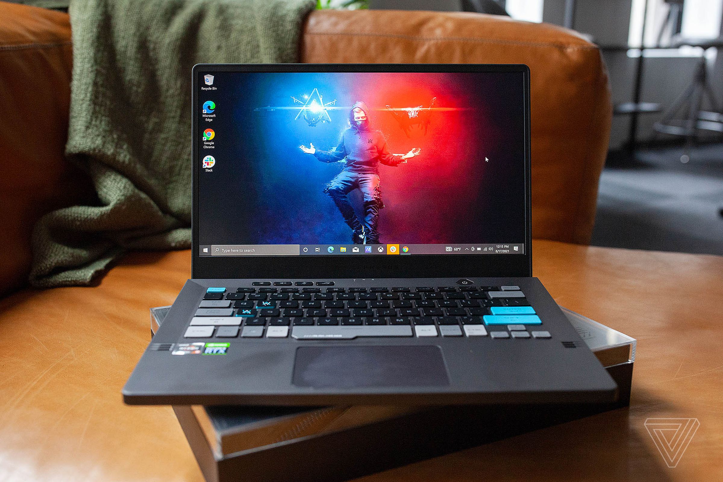 The Asus ROG Zephyrus G14 Alan Walker Edition on a couch seen from the front, open. The screen displays Alan Walker hovering on a red and blue background, conjuring lights of each color with his hands.