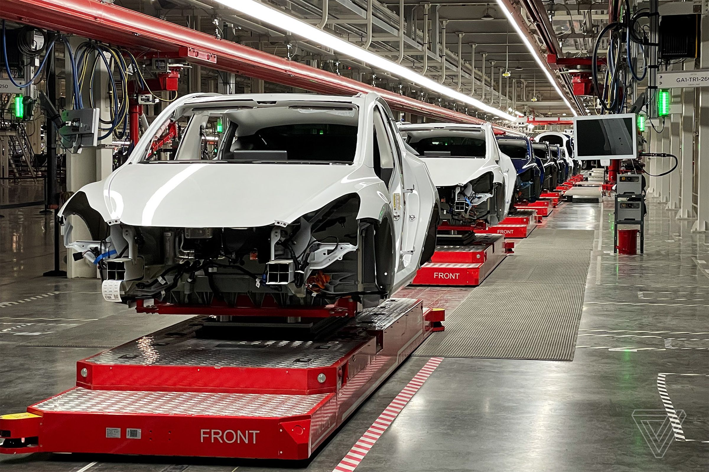 Unfinished Tesla model Y vehicles on factory assembly line platforms, no bumpers or headlights attached yet.