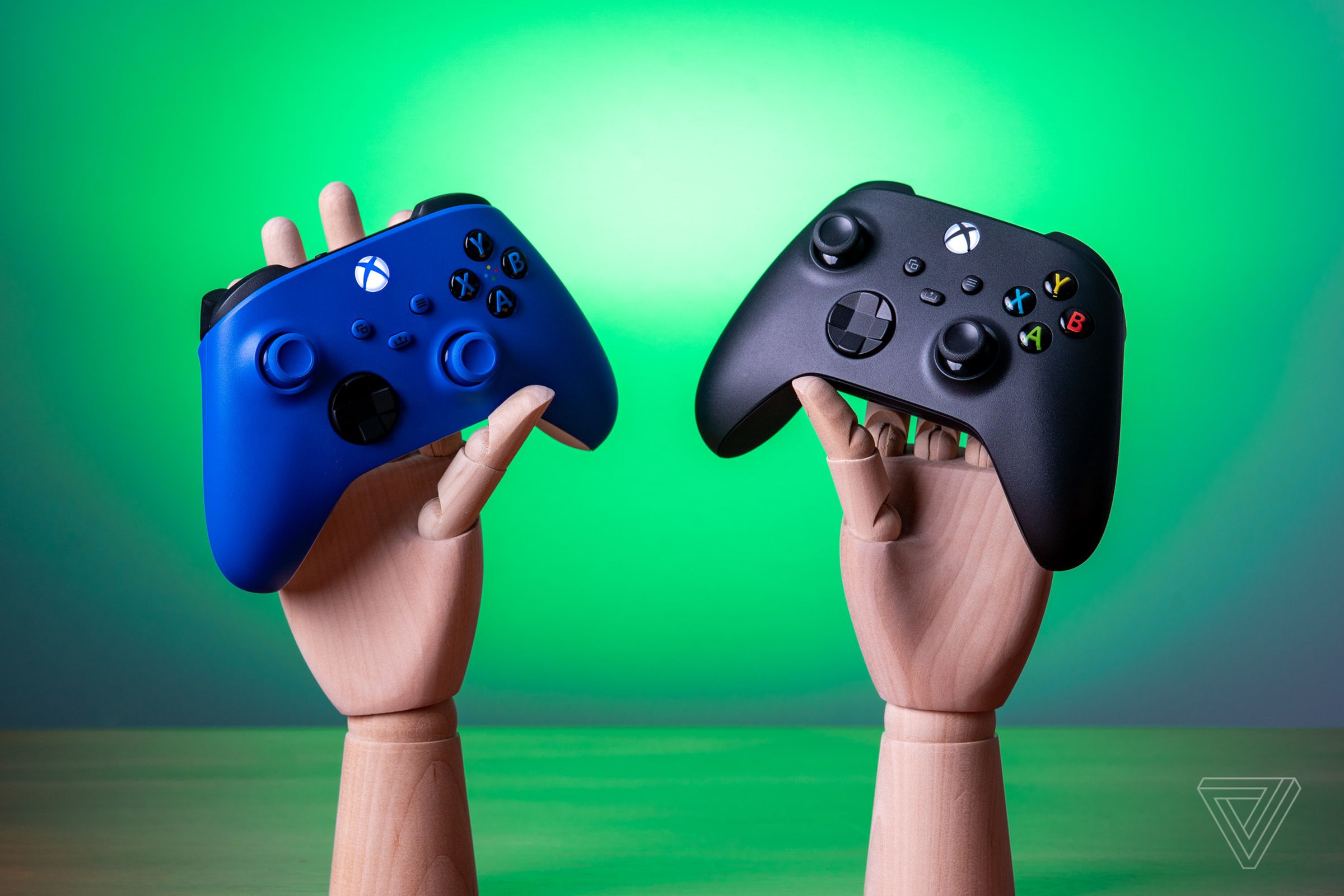 Two Xbox controllers, one blue and one black, being held by wooden mannequin hands.