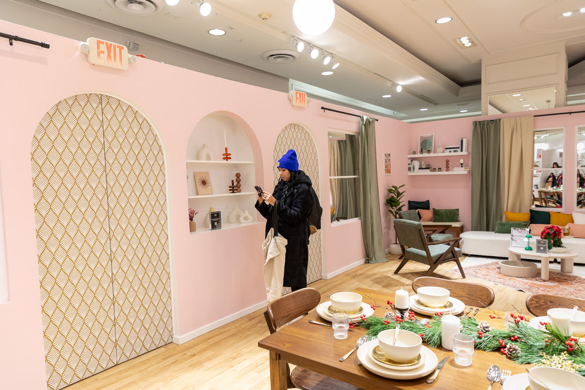 A constructed dining room featuring Shein products like dishes, decorations, and furniture. One shopper is walking through the space, looking at her phone.