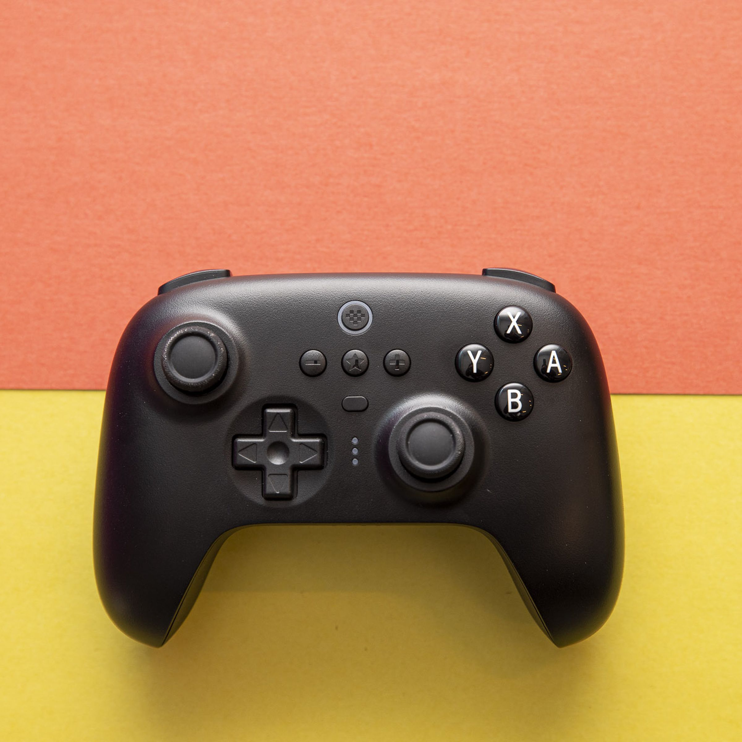 Top-down shot of 8BitDo Ultimate Controller on an orange and yellow background. The controller has an asymmetric joystick design, like an Xbox or Switch Pro controller, and it’s black, with white legends on the A B X Y buttons.