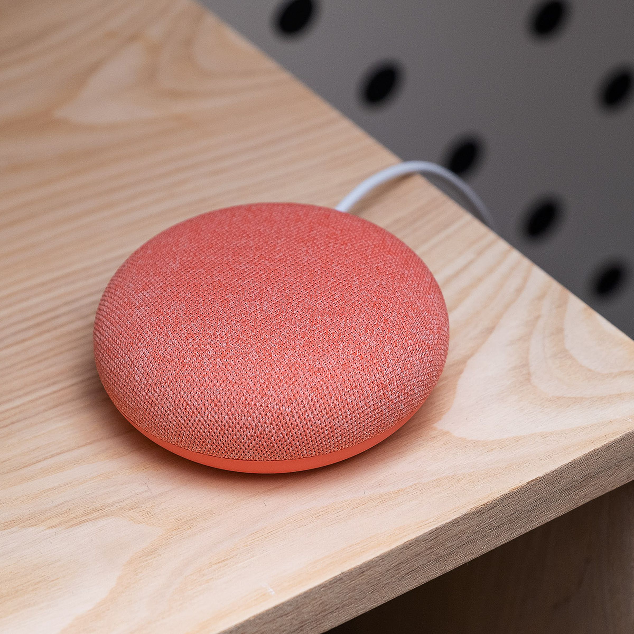 A coral colored Google Nest Mini smart speaker sitting on a wood desk.