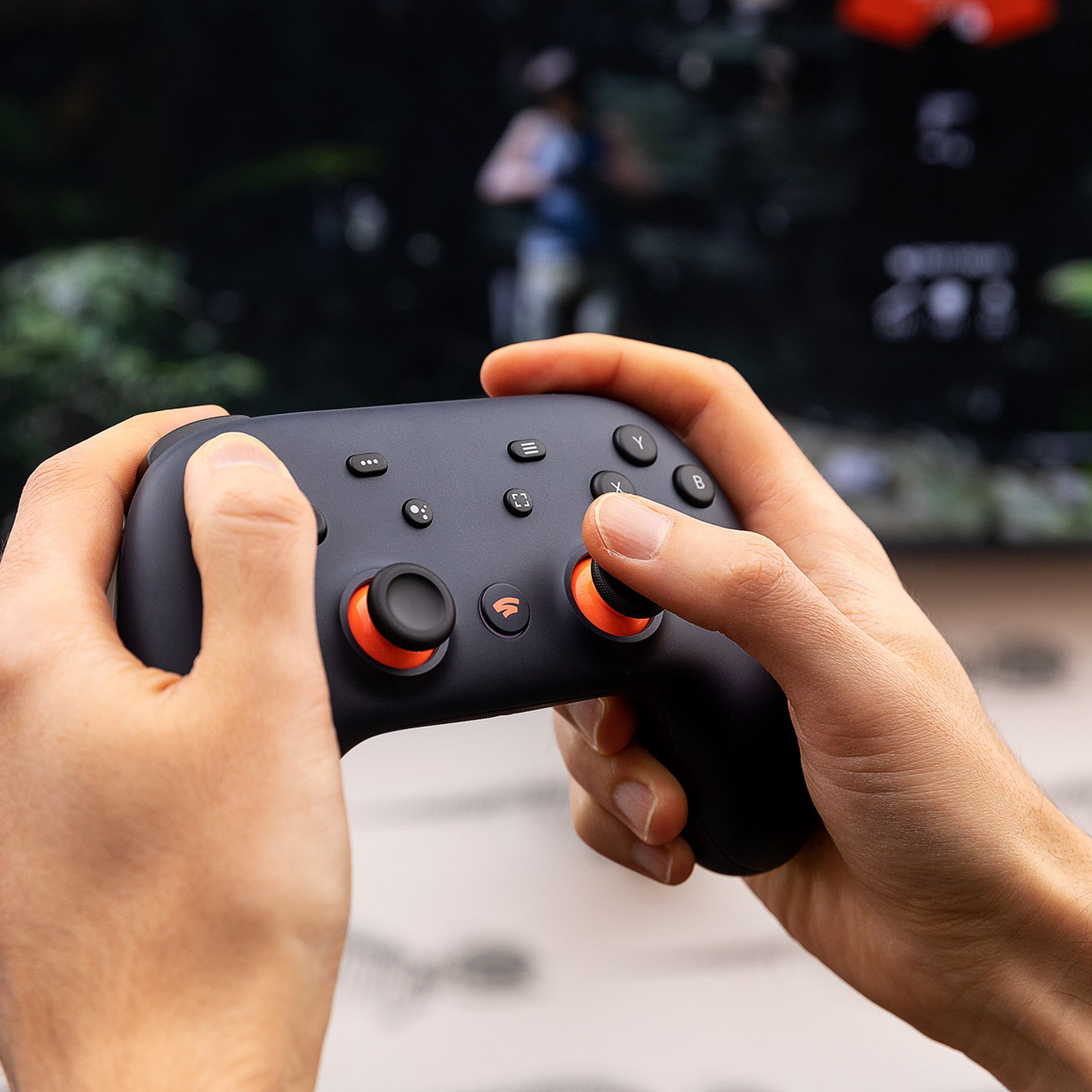 Two hands hold a Stadia controller in front of a TV.