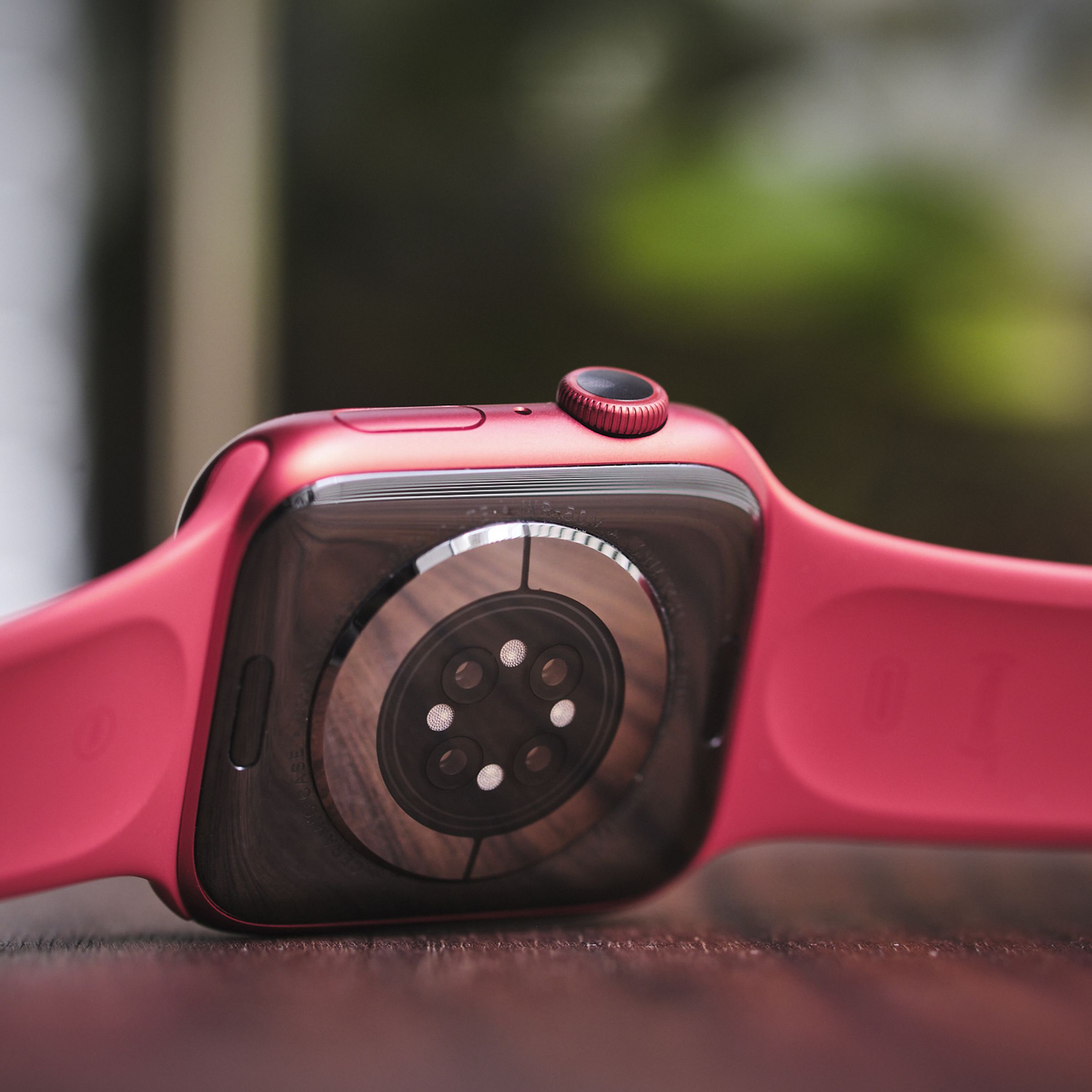 Close-up of rear side of an Apple Watch with a red sports band on a wood table, with its sensors showing