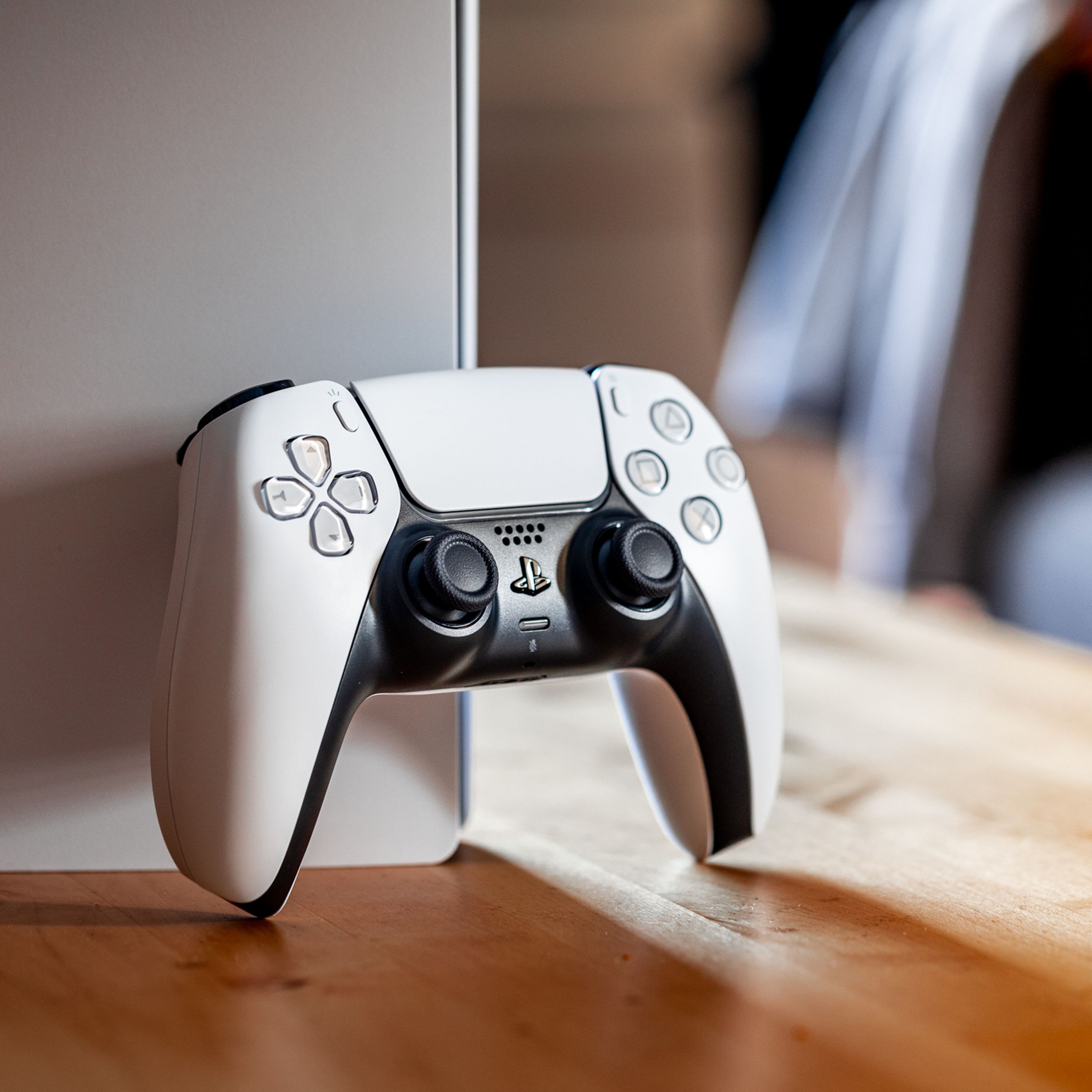 A white Sony DualSense controller resting against a PlayStation 5 console on a wood table.