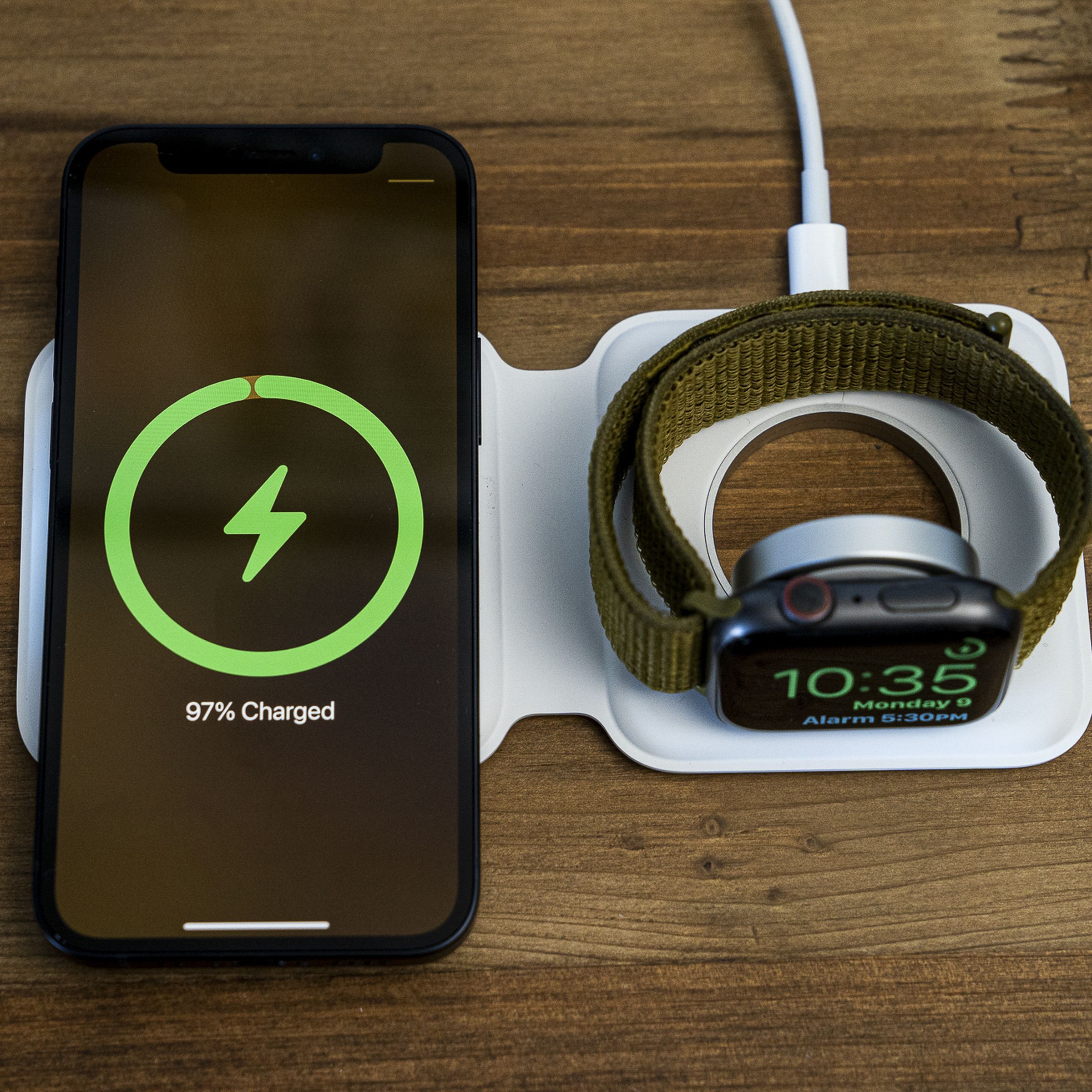 An iPhone and Apple Watch charging wirelessly on an Apple MagSafe Duo Charger, atop a wood table.