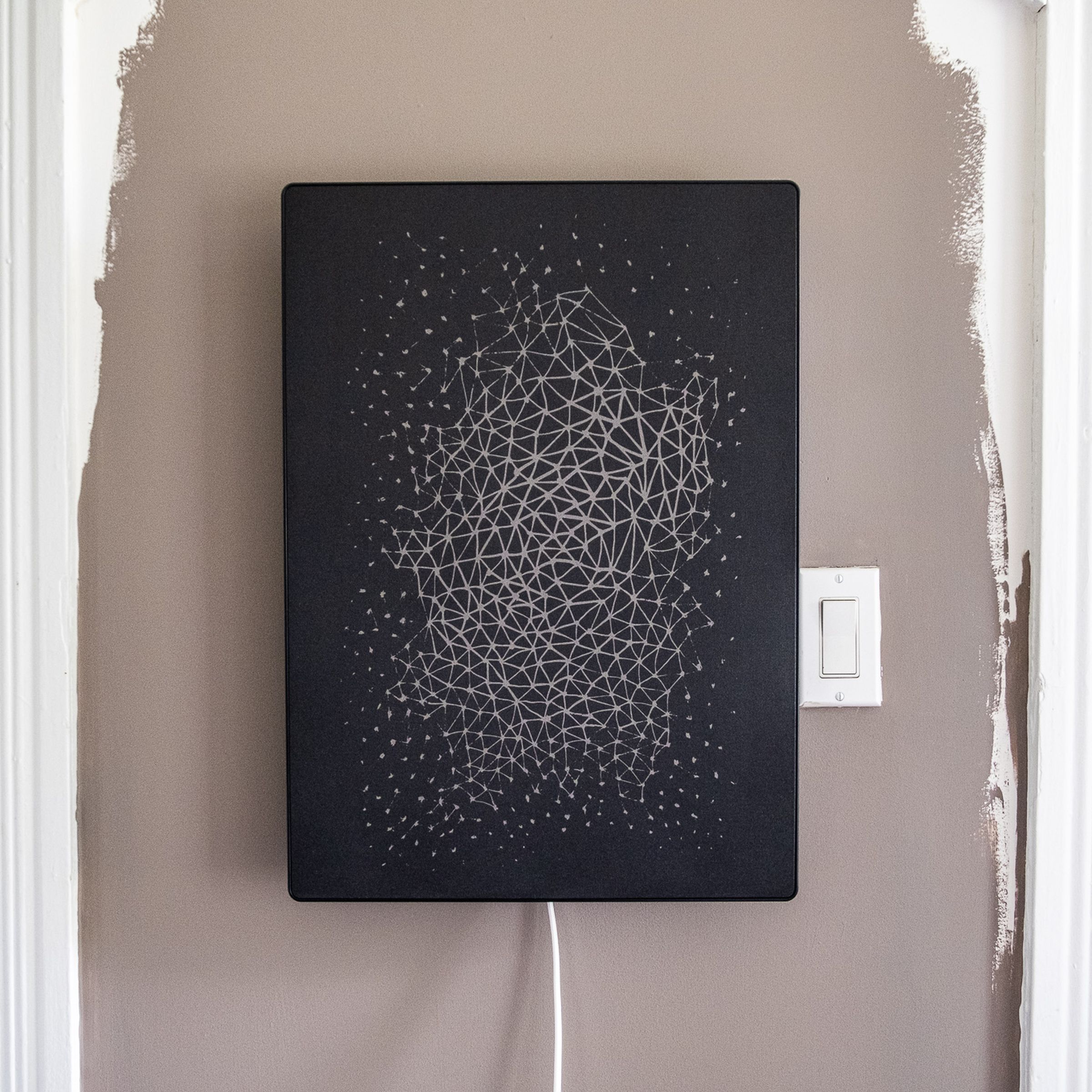 A black Ikea picture frame speaker with a geometric pattern design mounted on a wall beside a light switch.
