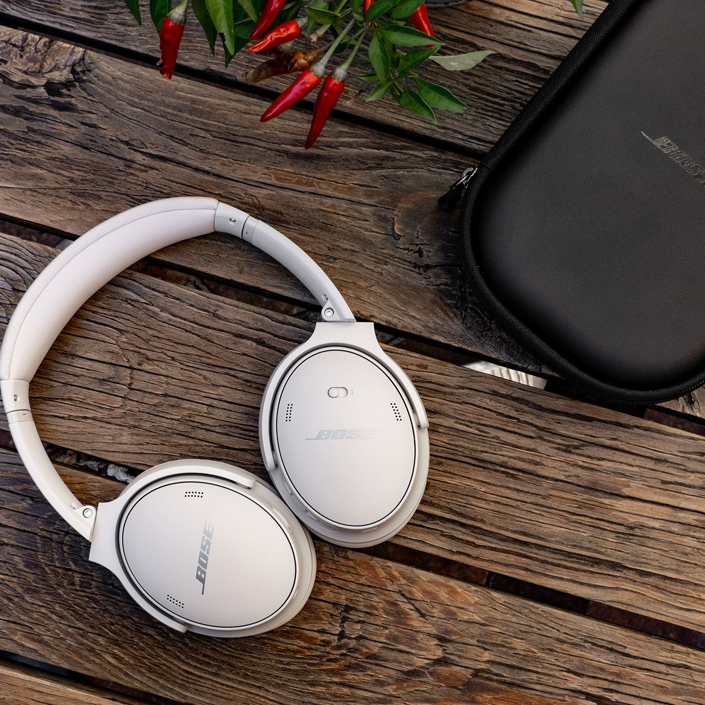 The white version of the Bose QuietComfort 45 headphones on a wood picnic table with their black protective zipper case beside them.