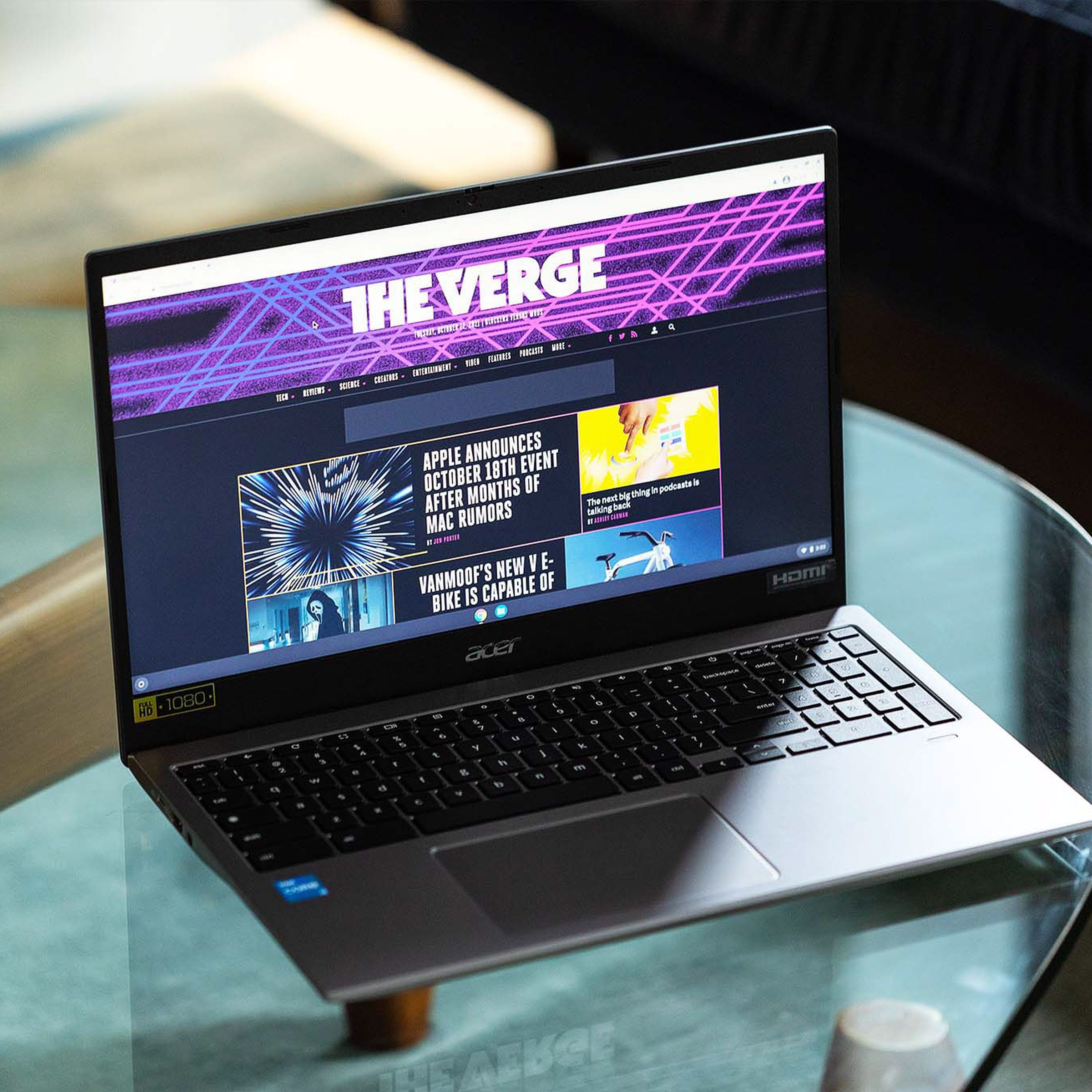 The Acer Chromebook 515 open, to the right, on a clear table. The screen displays The Verge homepage.