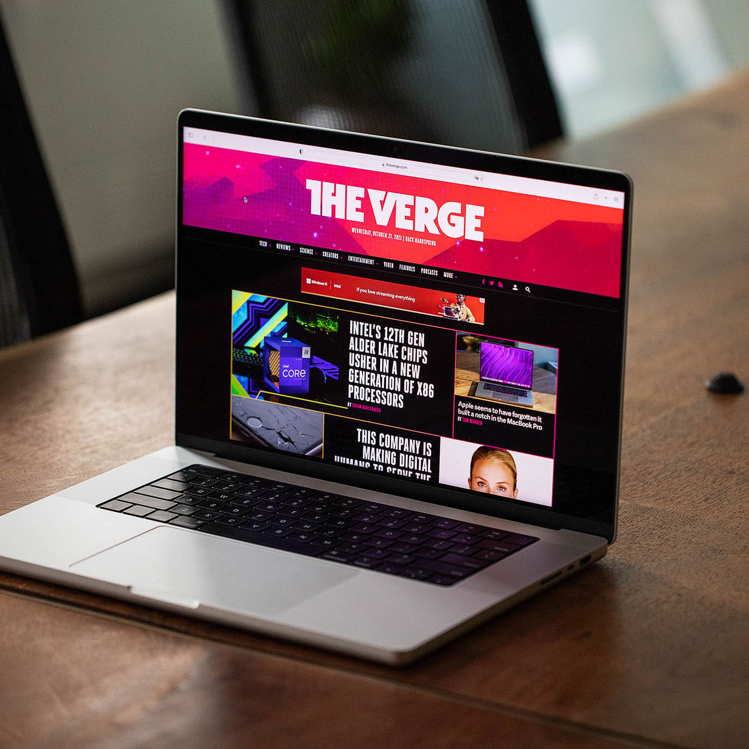 Apple’s 2021 14-inch MacBook Pro is opened on a wooden table. Its display is showing off The Verge’s homepage.