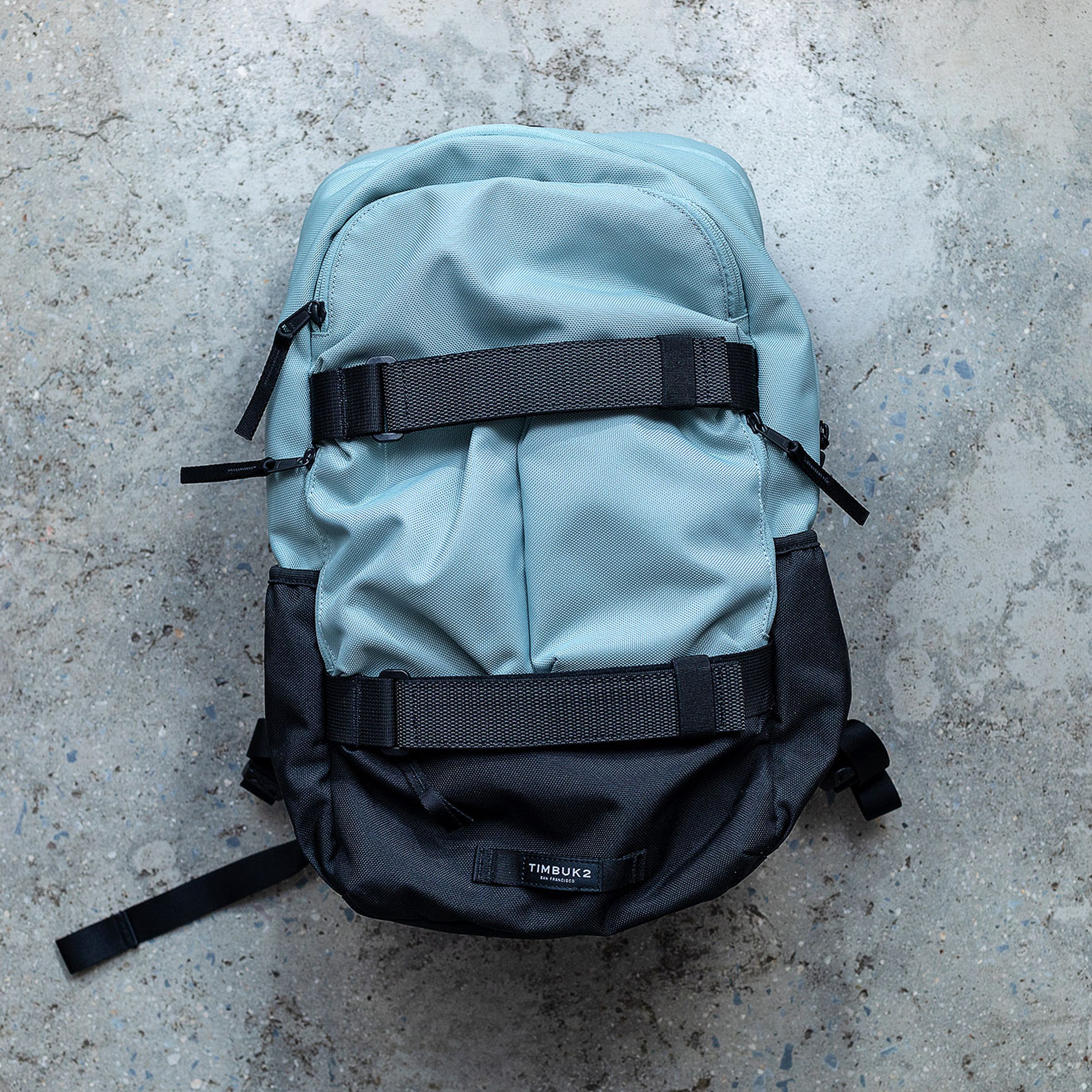 A two-tone light-blue / black backpack laying on a gray concrete floor, with its rear side pointing upwards toward the camera.