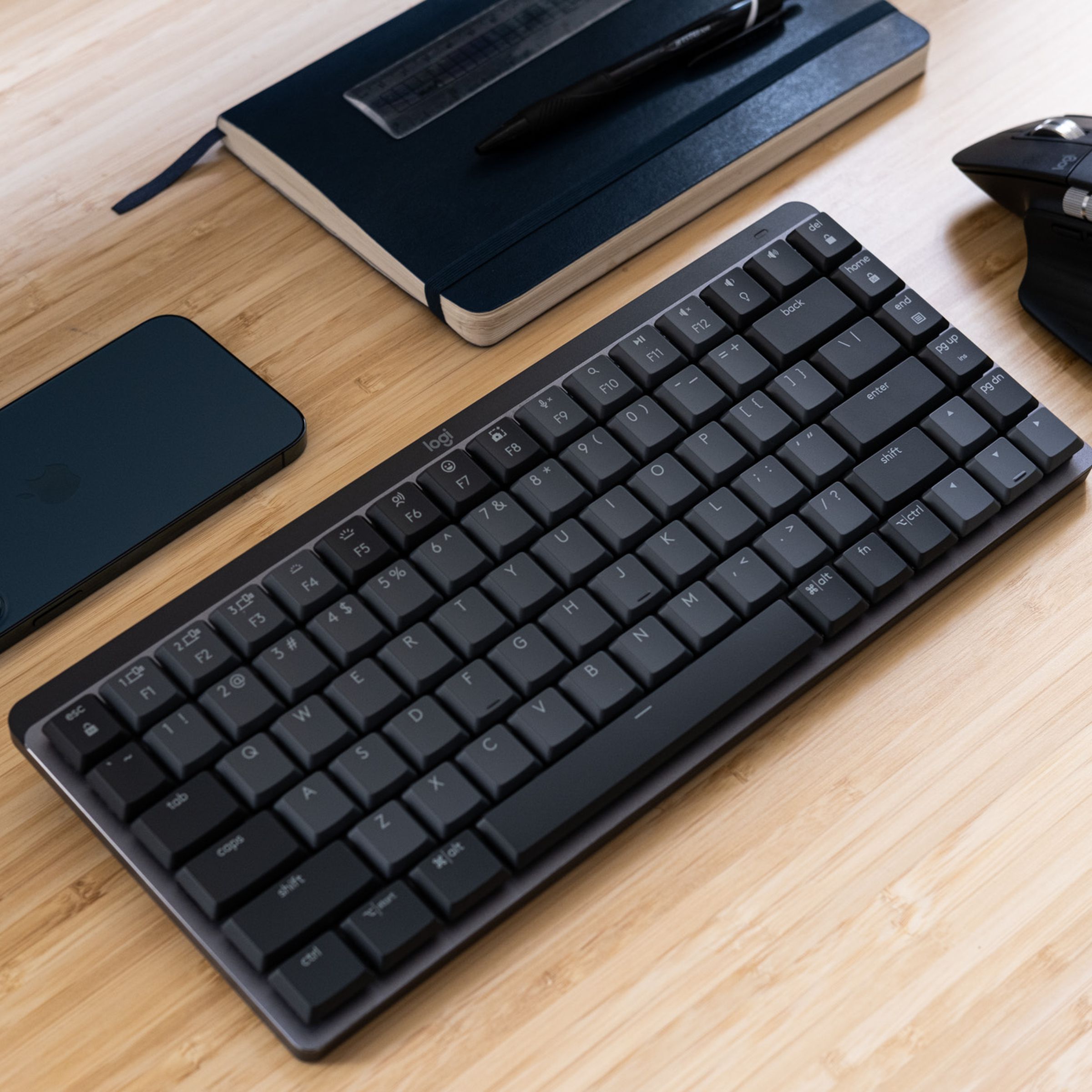 A Logitech MX Mechanical Mini keyboard on a wood desk, beside a mouse, phone, and notebook.