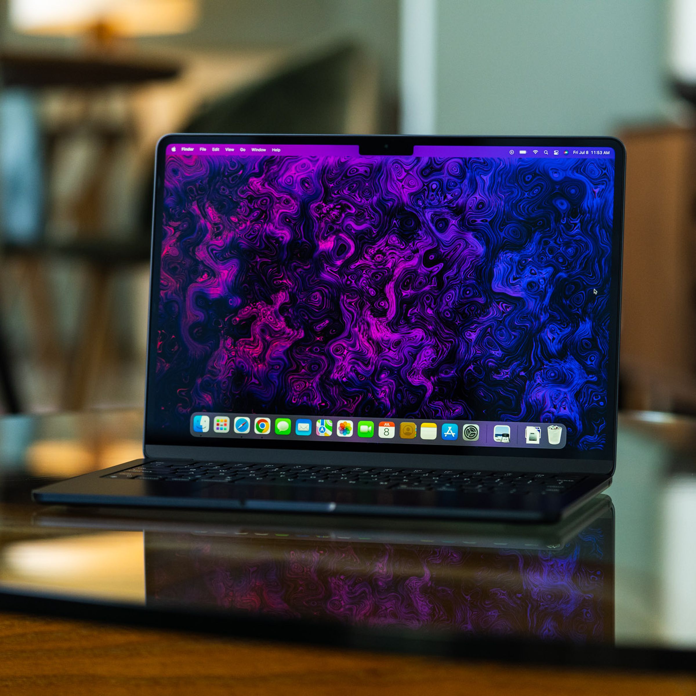 El MacBook Air M2 está abierto, frente a la cámara.  Su pantalla está iluminada y muestra un fondo psicodélico morado y negro creado por el equipo de arte e ilustración de The Verge.