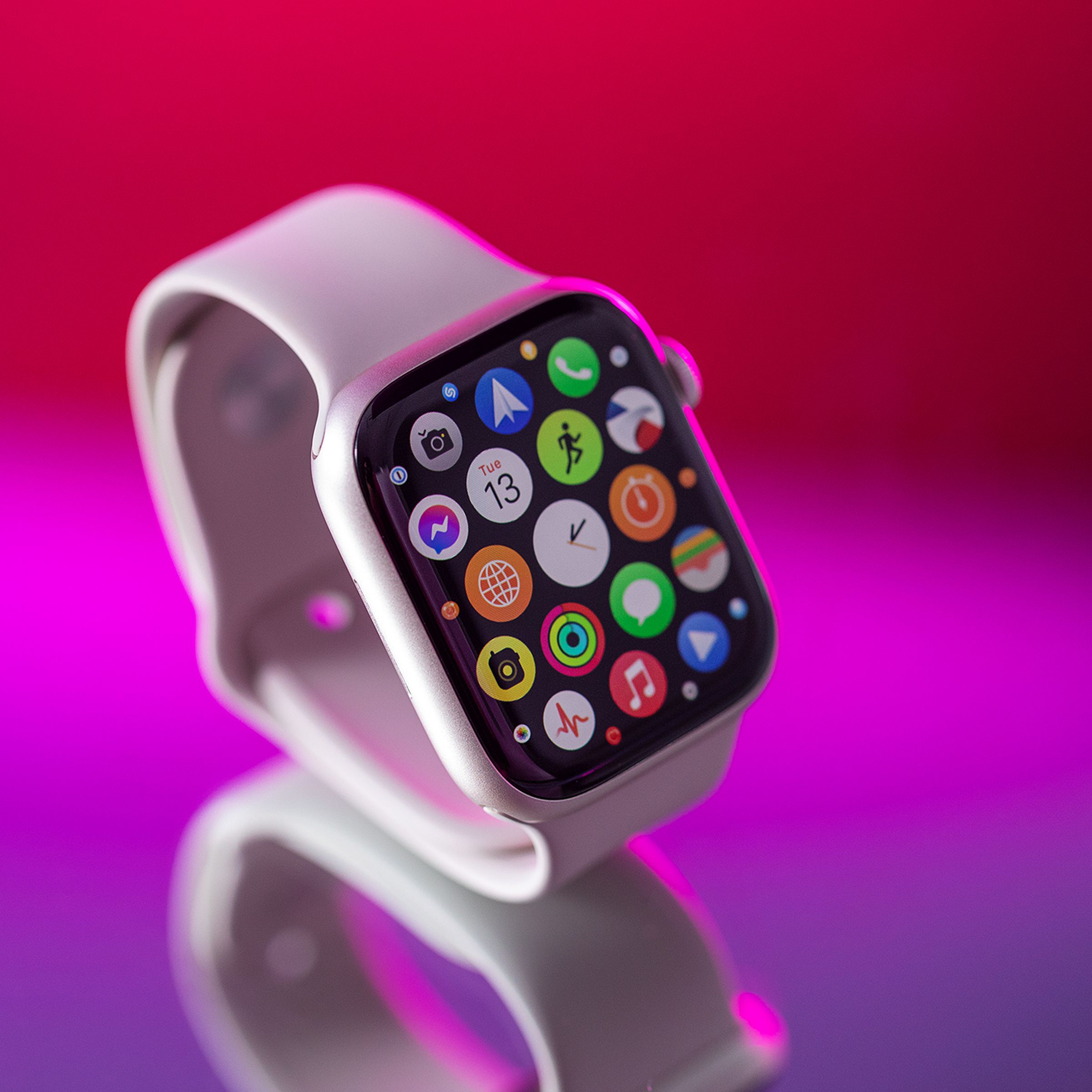 Close-up photo of a silver Apple Watch Series 8, sitting atop a reflective tabletop surface, highlighted by a magenta backlight. The screen of the watch is on the watch OS app grid home screen.