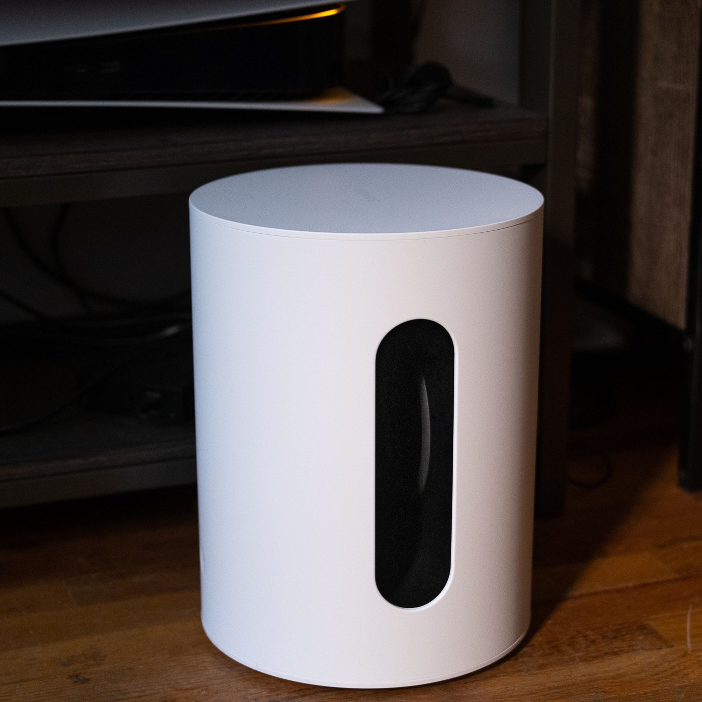 An image of the Sonos Sub Mini pictured on the floor with a TV stand and various components behind it.